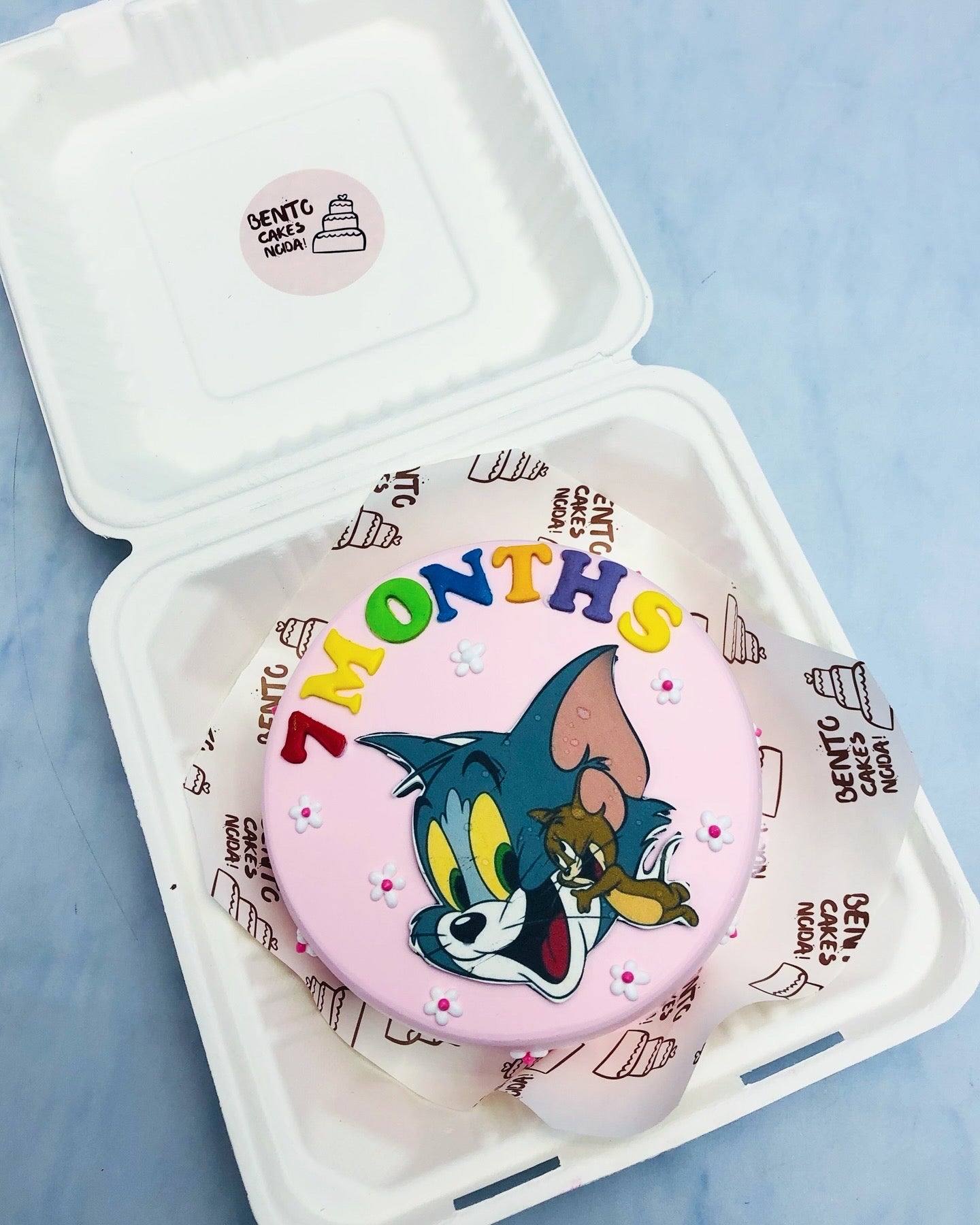 A light pink cake with a Tom and Jerry cartoon print and a text  written with colorful fondant alphabet.