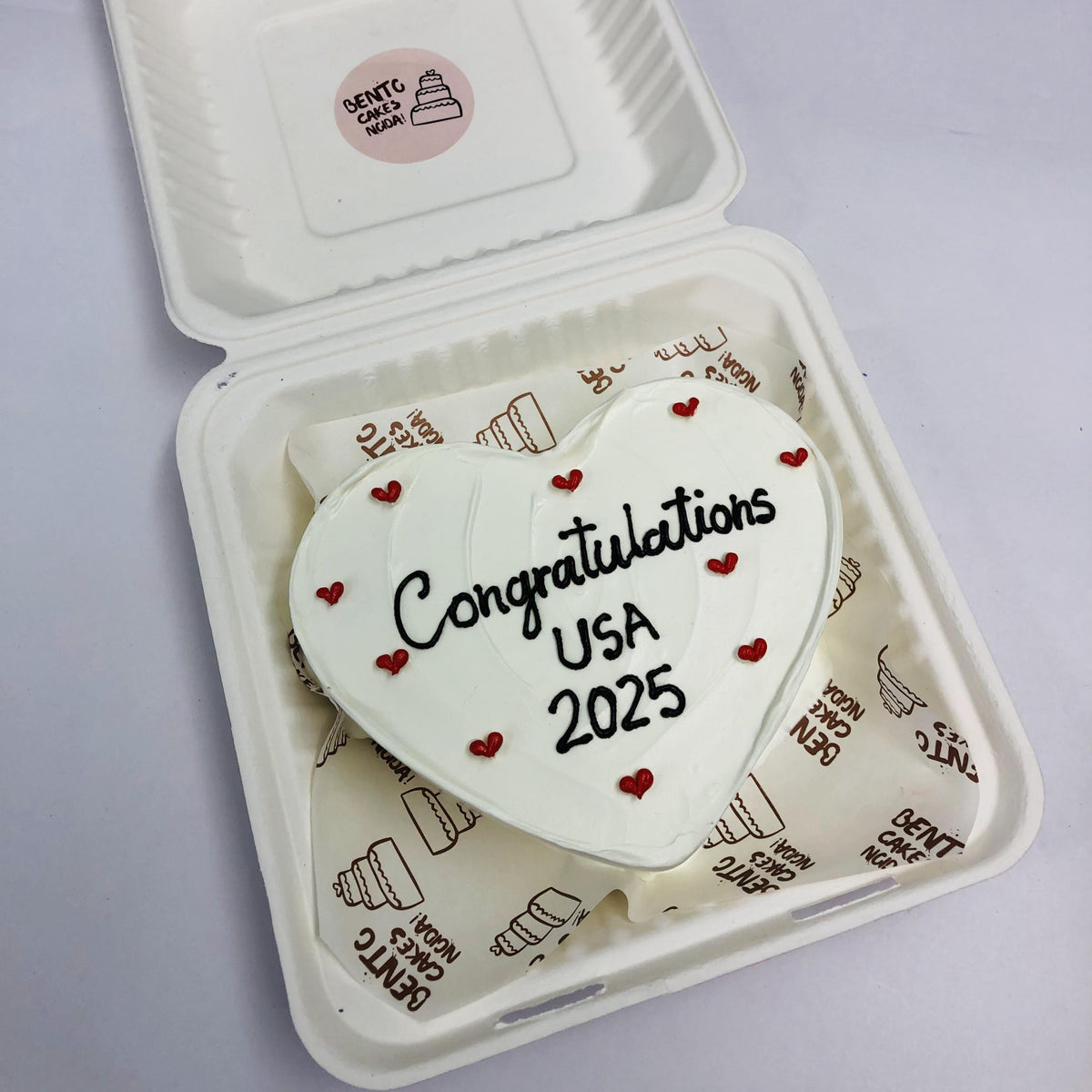 A white heart shape cake filled wih red hearts and wavy design on it with "Congratulations USA 2025" text on it.