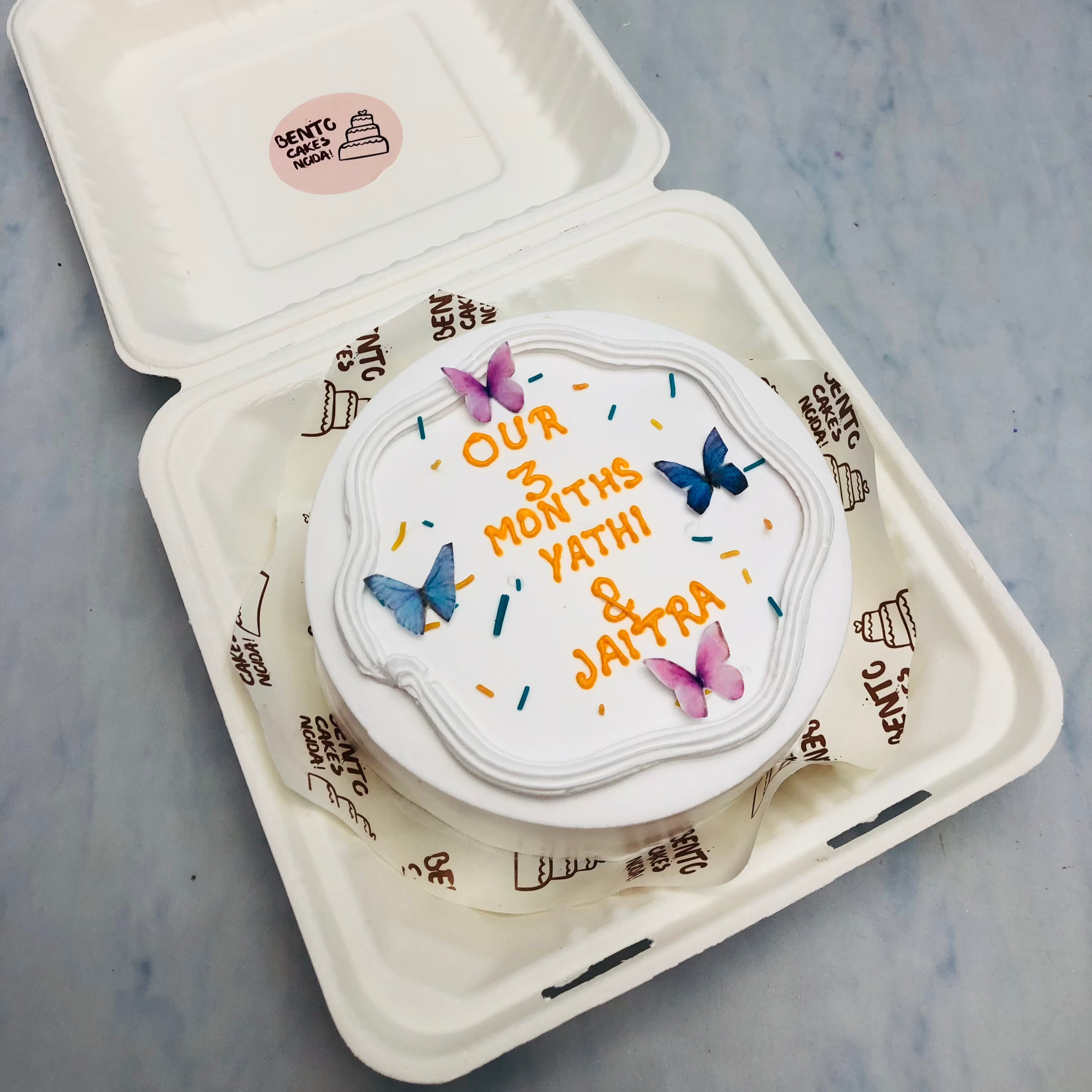 A white bento cake with blue and pink butterflies and a cute text written with orange color.