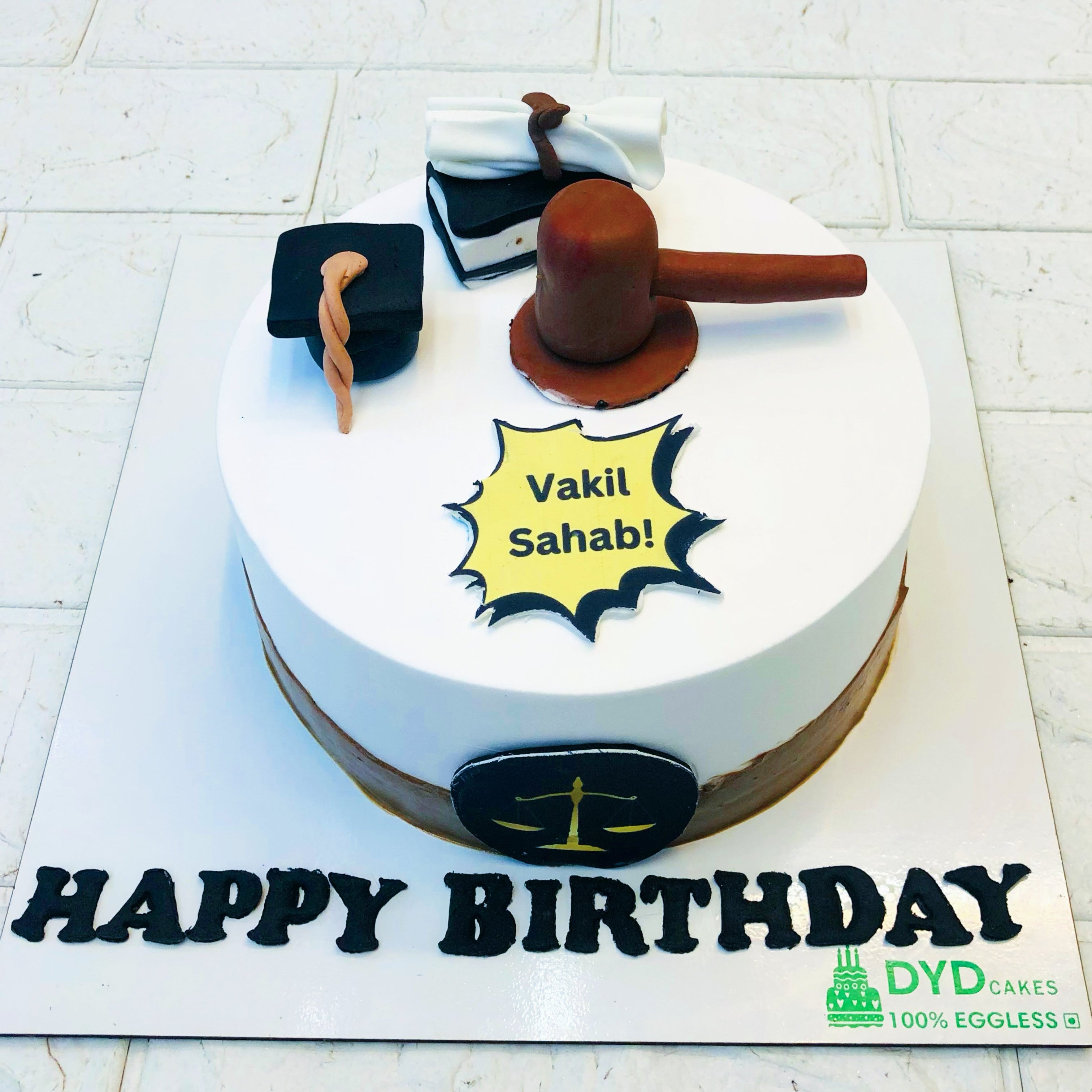 A white and brown cake is designed on lawyer theme having a cap, book and court hammer on its top and a "HAPPY BIRTHDAY" written on front.