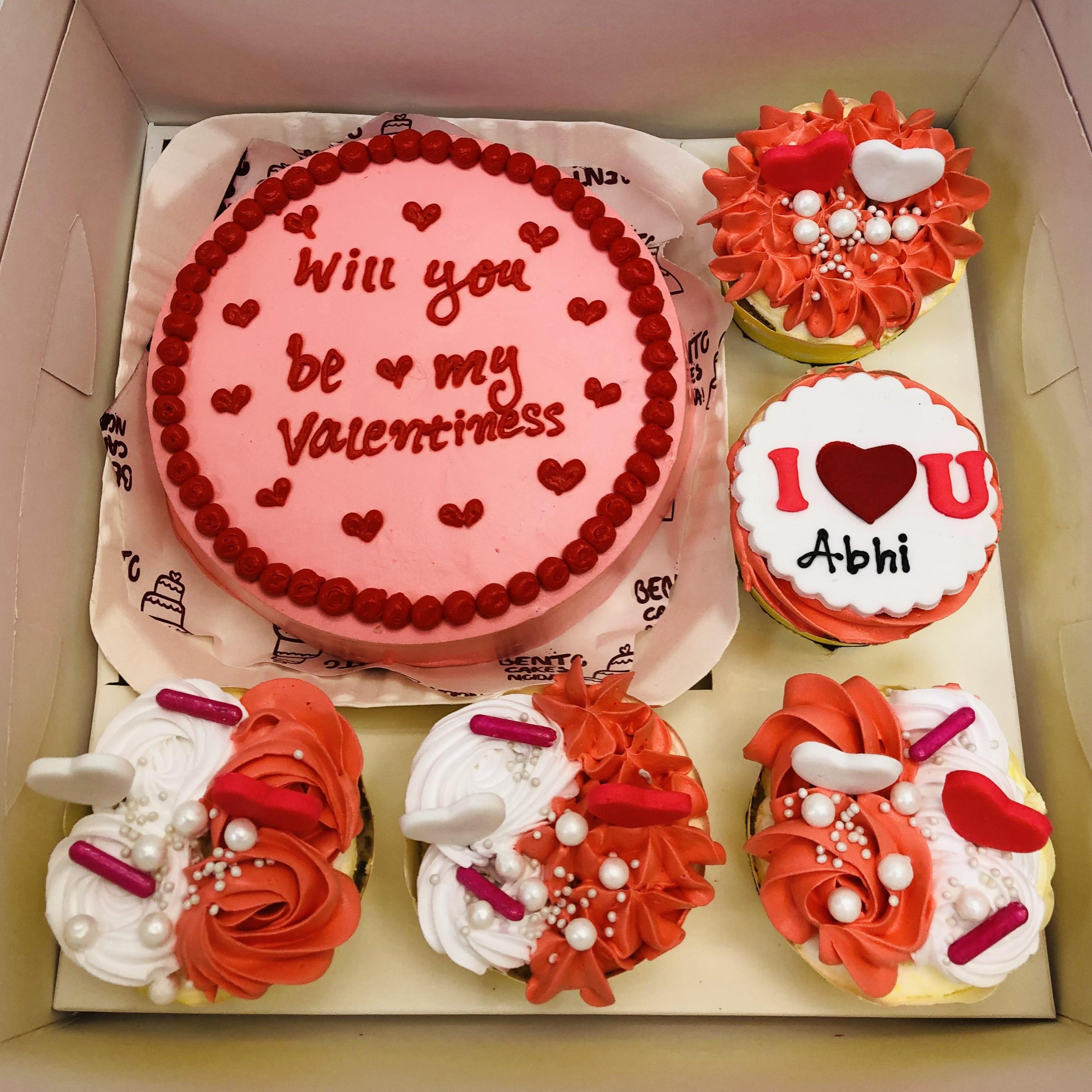 A Valentine's themed bento combo cake set featuring a round pink cake with red decorations and the text "Will you be my Valentine" written on it. Surrounding the cake are four cupcakes decorated in romantic red, white, and pink frosting with heart-shaped toppers and pearl-like sprinkles. One of the cupcakes includes the message "I ❤️ U Abhi."