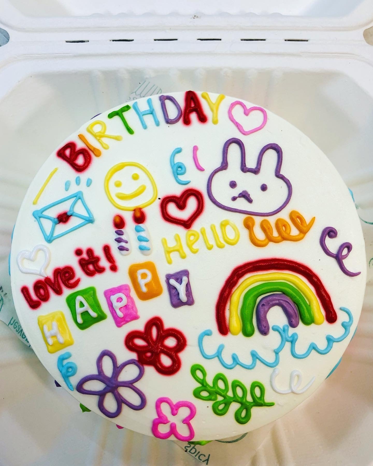A colorful birthday cake with various decorations including a rainbow, flowers, a bunny, and cheerful messages.