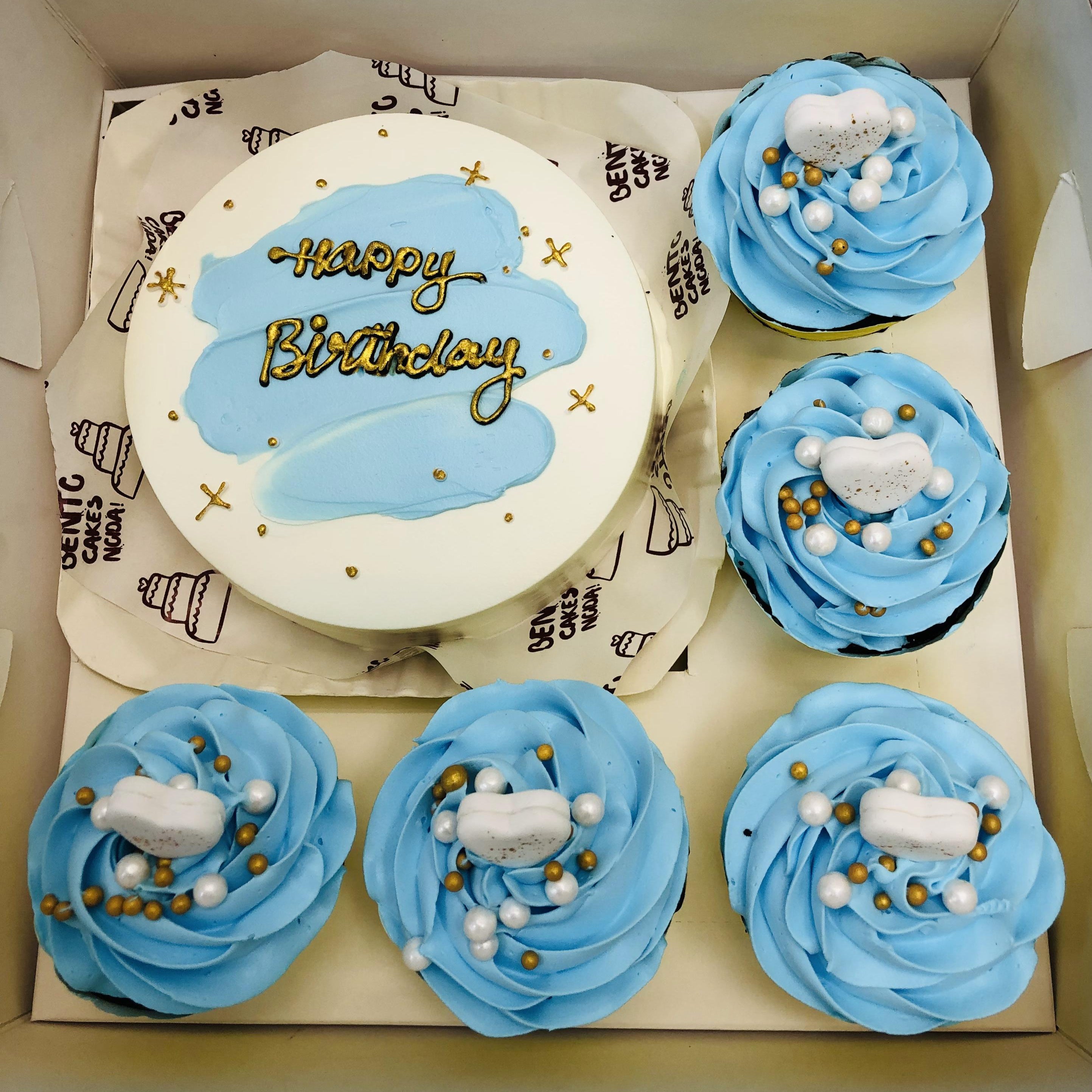 A boxed set featuring a round cake with white frosting and light blue brushstroke decorations. The cake has the text "Happy Birthday" written in gold. It is accompanied by five cupcakes with light blue frosting, topped with white and gold decorative elements, such as small white and gold balls and heart-shaped toppers.