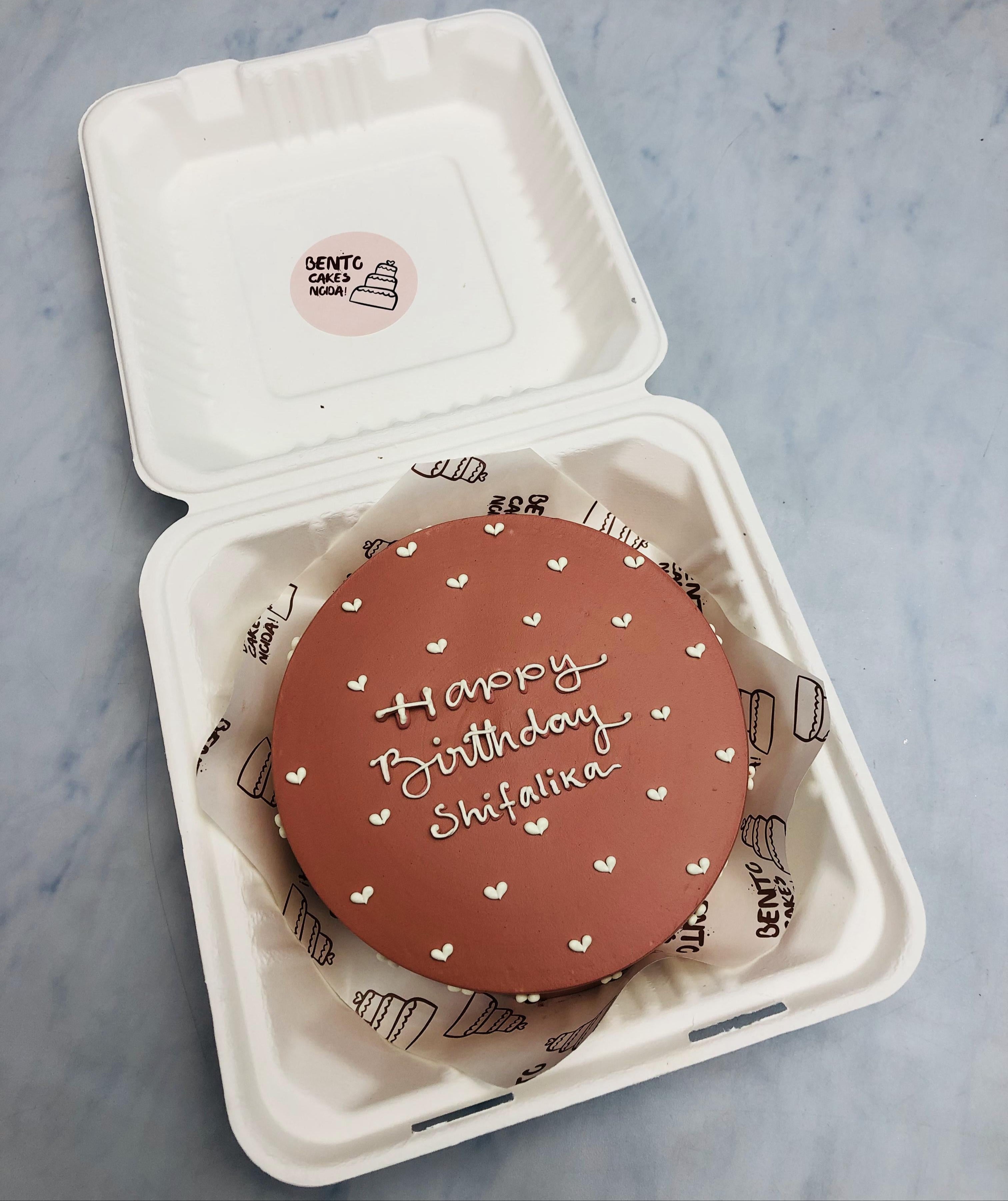 A round, brown birthday cake with white icing that reads "Happy Birthday Shifaika" is placed inside an open white biodegradable container.