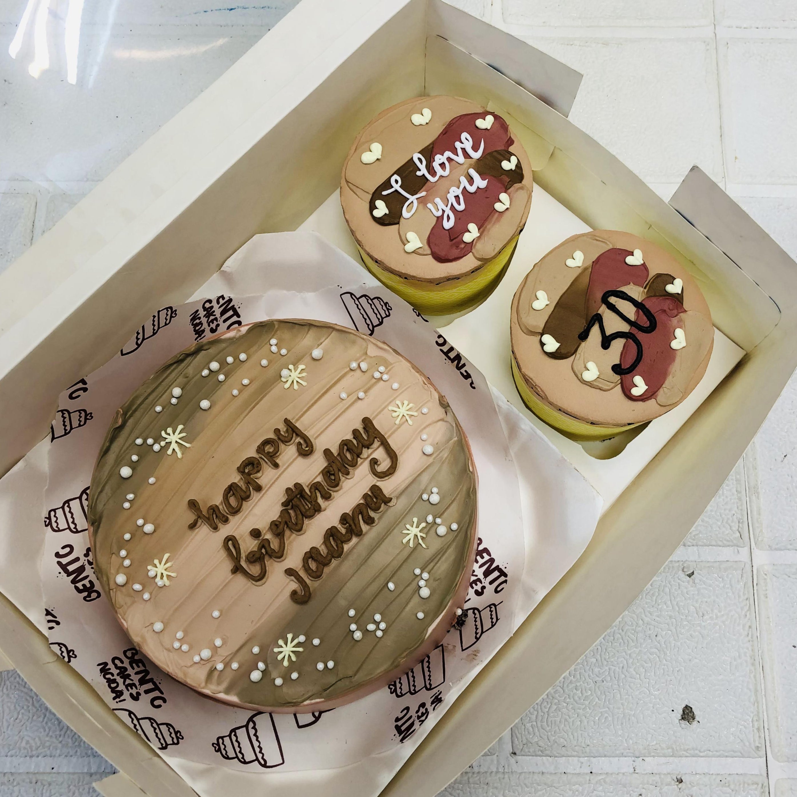 A boxed set featuring a gradient brown mini bento cake with white and yellow floral decorations and the message "Happy Birthday Joann" in white icing. Accompanied by two cupcakes: one with "I love you" and heart decorations, and another with "30" in white icing and hearts.