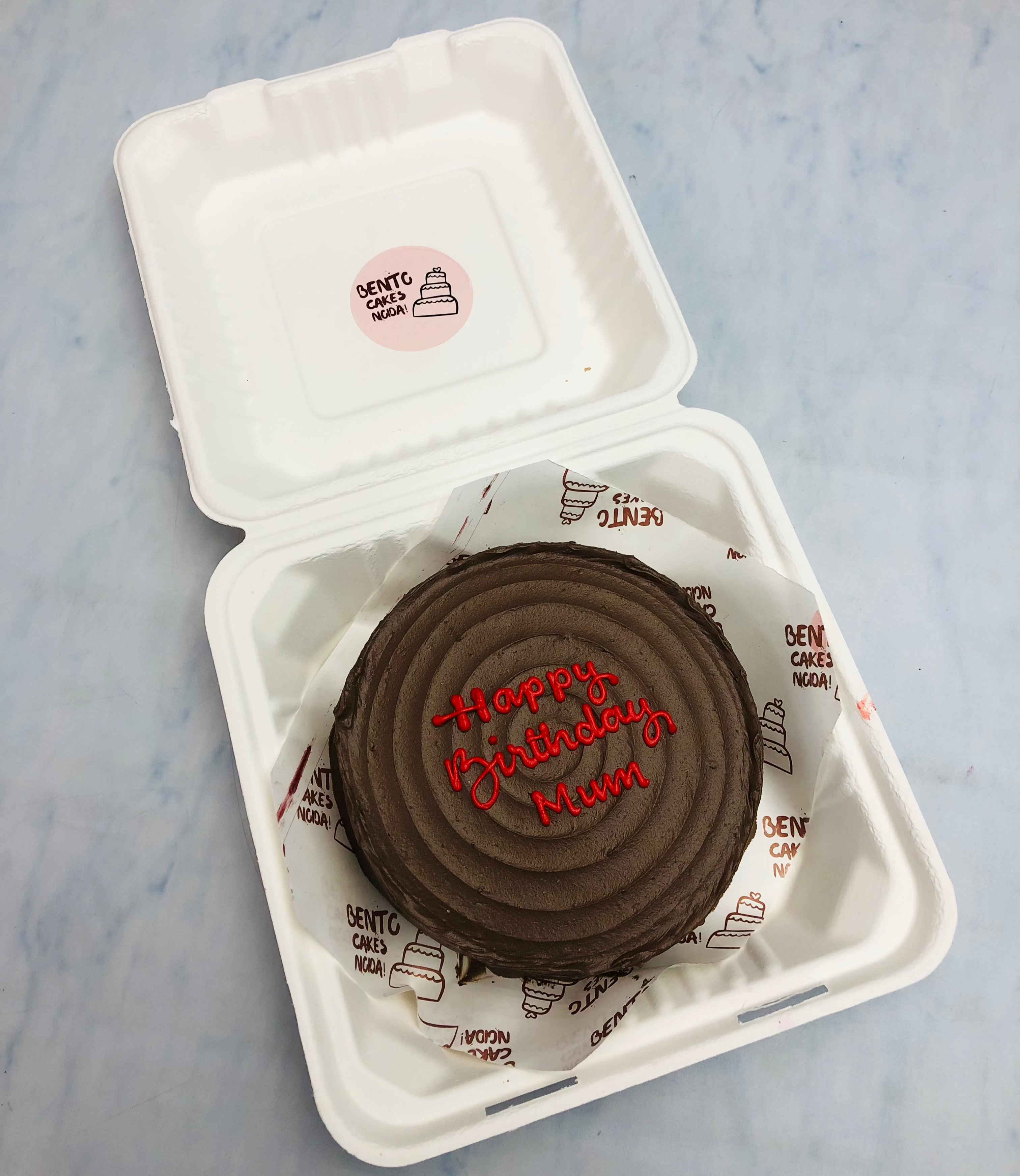 A chocolate brown bento cake having  wavy desgn on top with "Happy Birthday Mum" text written with red icing.