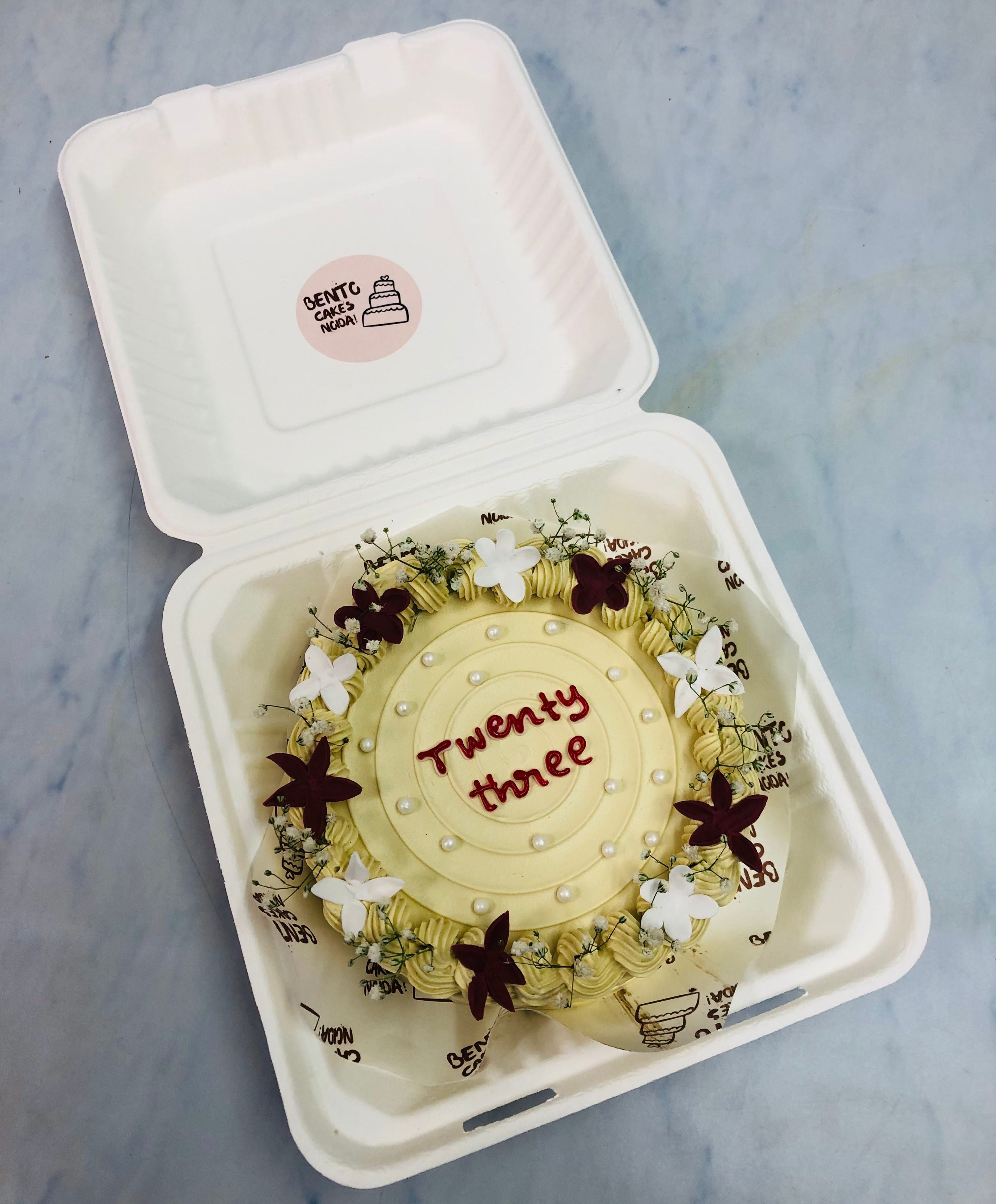 A beautifully decorated birthday cake inside a white bento box. The cake features delicate white and dark red floral designs, small white pearls, and the words "Twenty three" written in red icing on top.