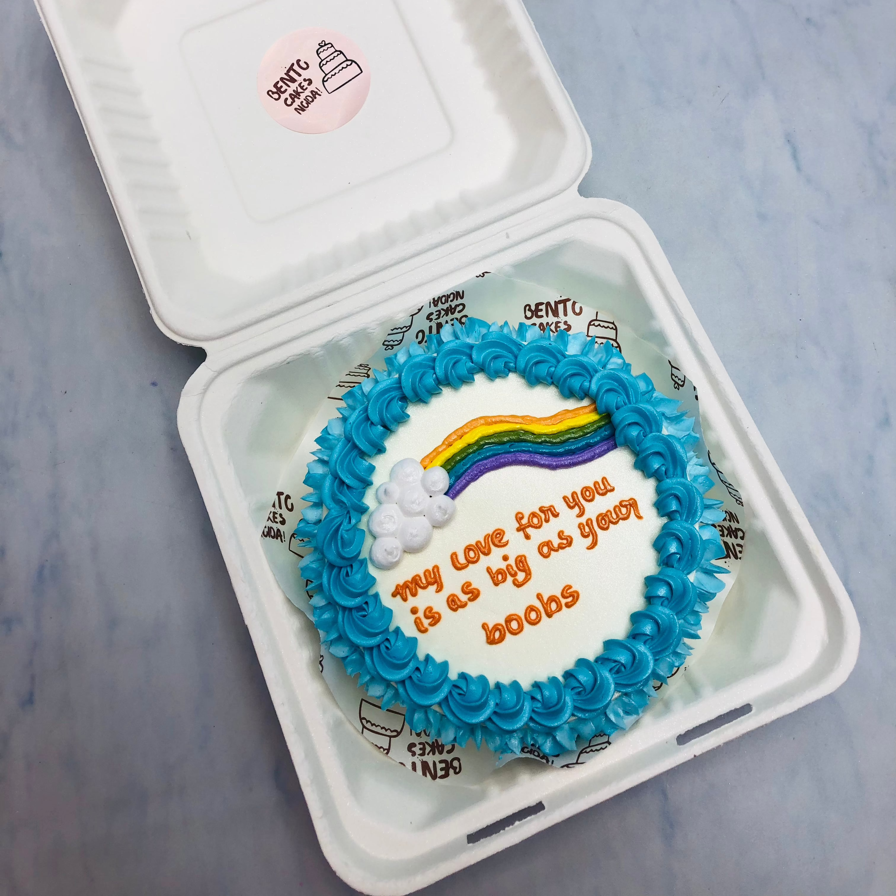 A white bento cake with blue truffle boundary and rainbow design in center with a vulgar text.