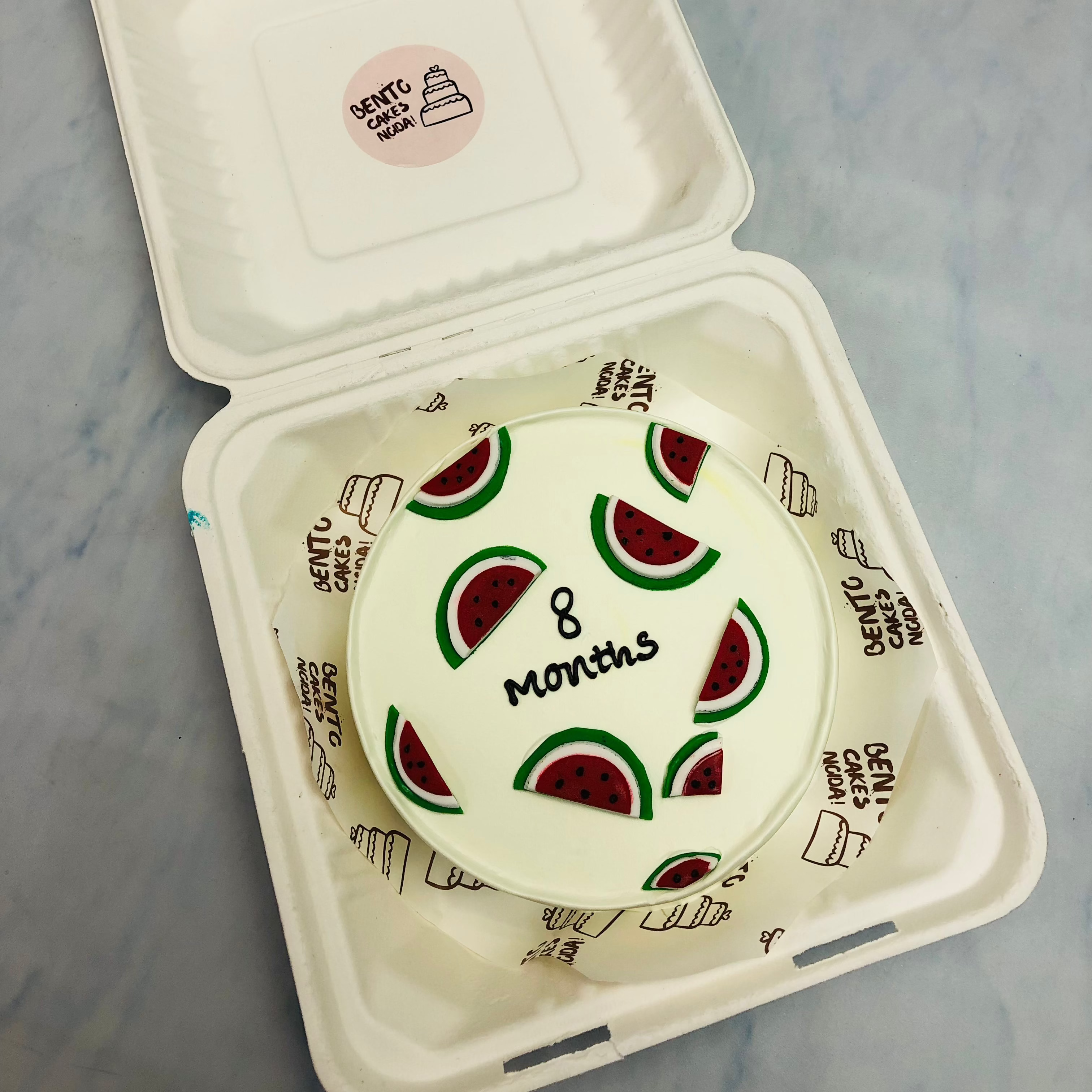 A white bento cake with watermelon pieces design all over the cake with "8 Months" text.