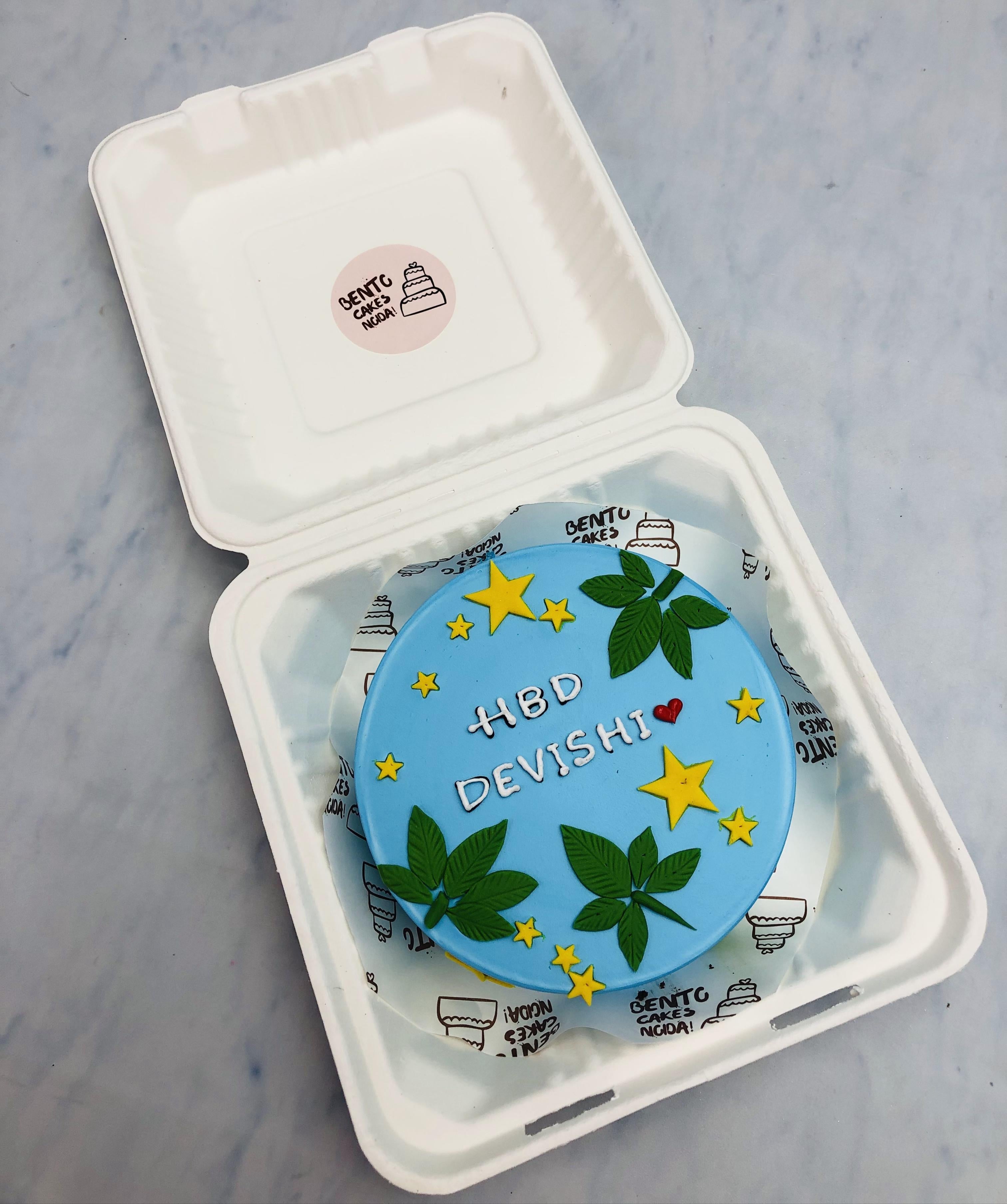 A compact blue bento cake designed for a birthday celebration, featuring 'HBD DEVISHI' in yellow letters, accompanied by green leaf and yellow star decorations inside a bento box.