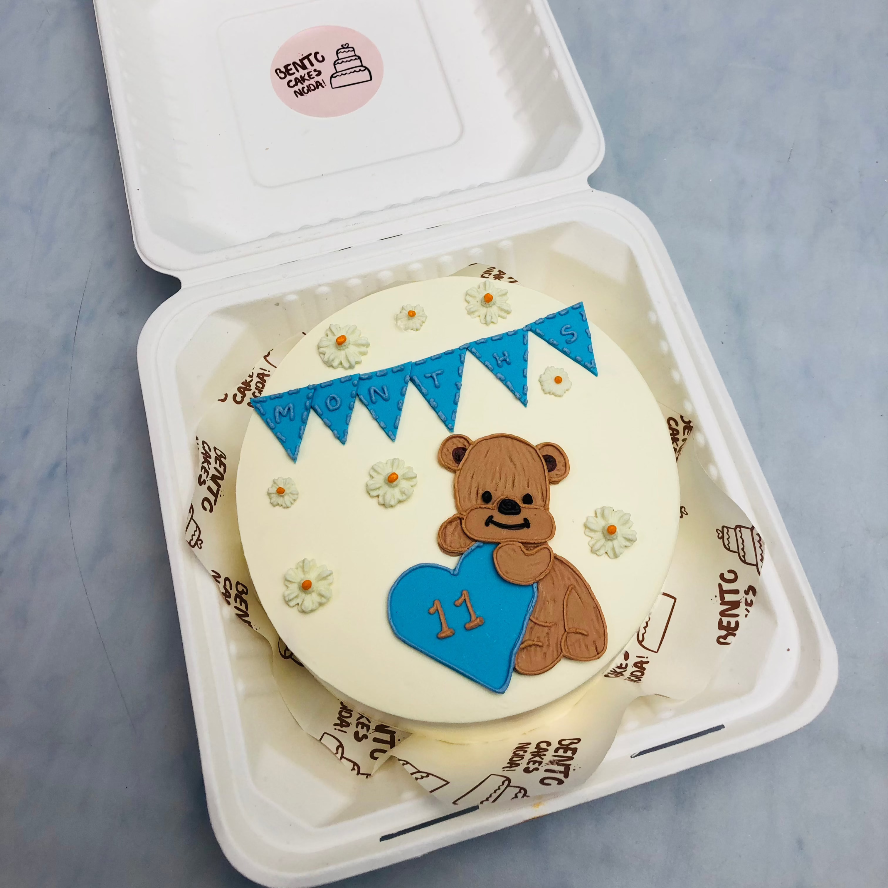 An offwhite bento cake with a brown teddy bear taken 11 number tag and cake filled with floral design.