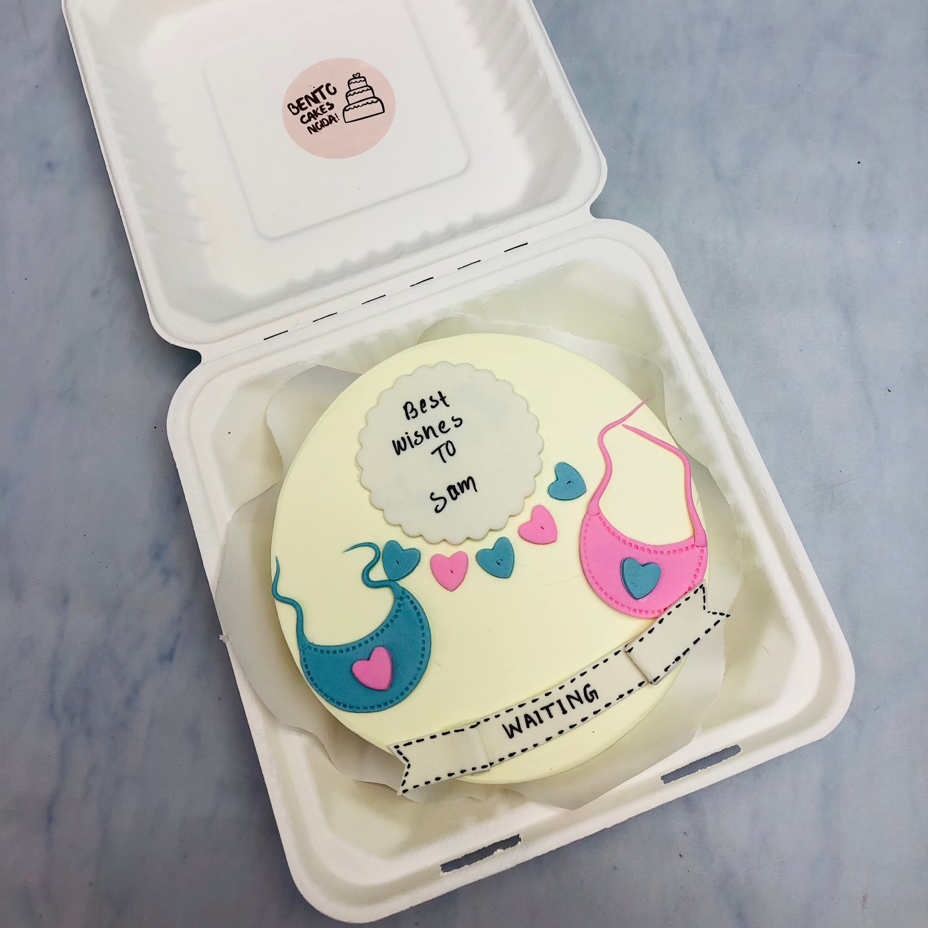 An offwhite cake in a bento box with coming baby desses design in blue and pink fondant.