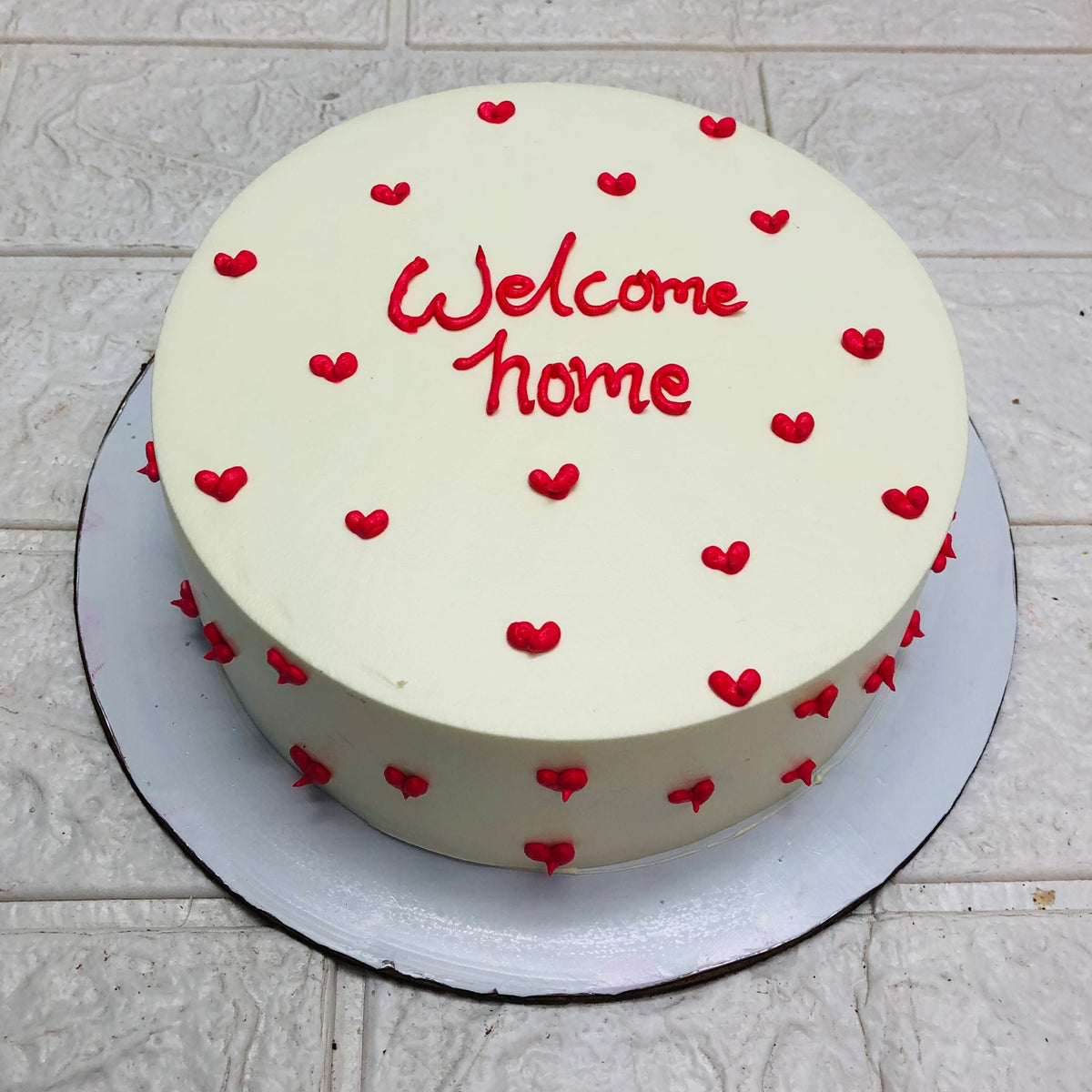 A round white cake with red heart decorations and the text "Welcome Home" written in red icing on top. The cake is placed on a white cake board, set against a tiled backdrop.