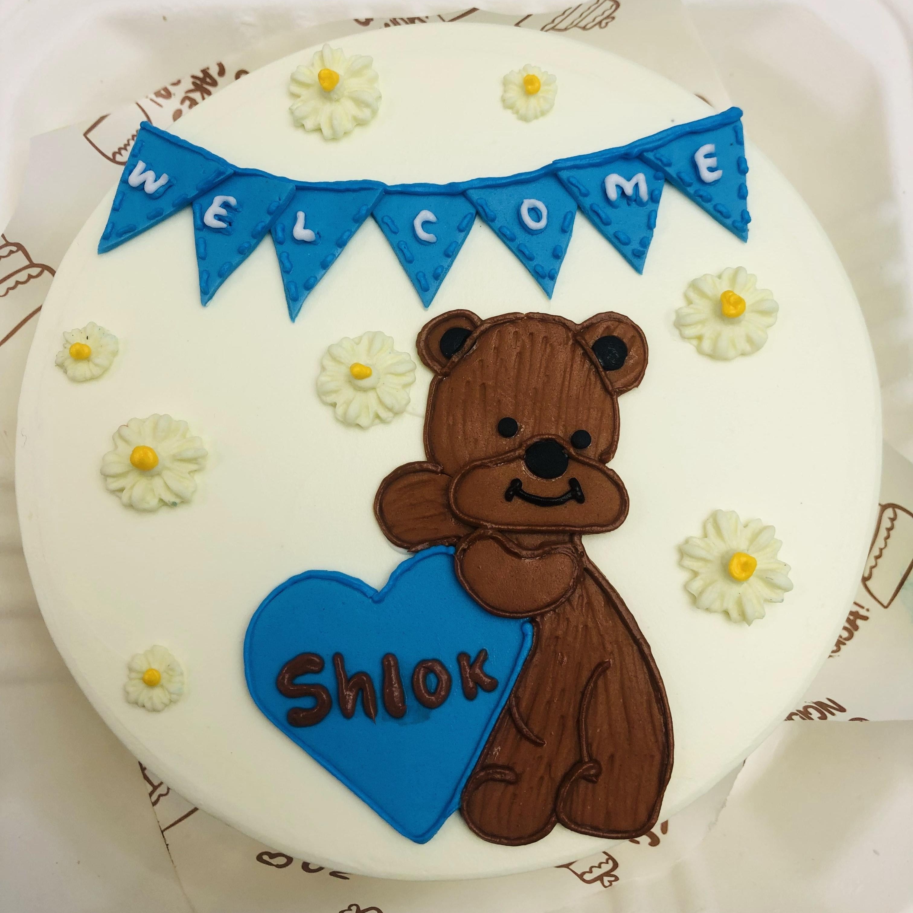 A white bento cake with white floral design and a tedy bear welcoming "Shlok"