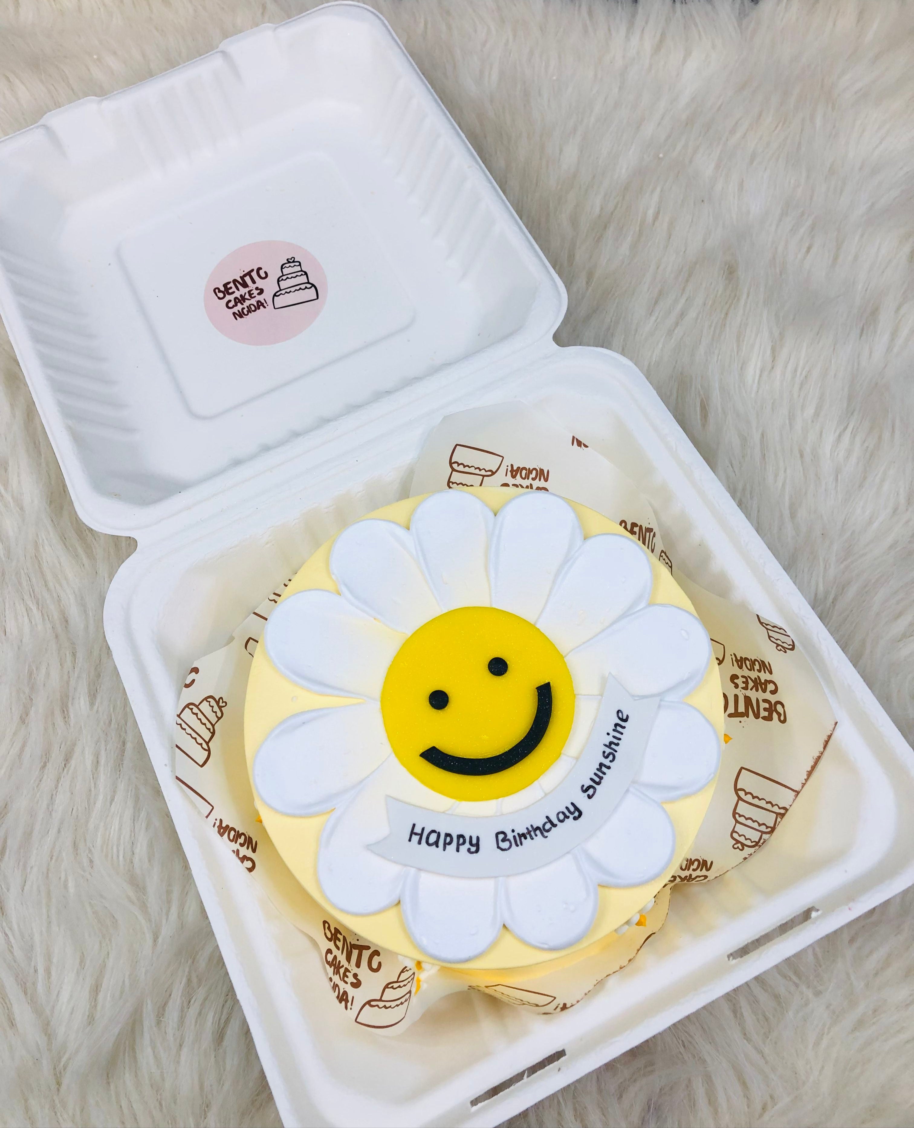 A delightful bento cake in a white takeout container, decorated to resemble a cheerful daisy with white petals and a yellow center featuring a smiling face. The cake includes the message "Happy Birthday Sunshine".