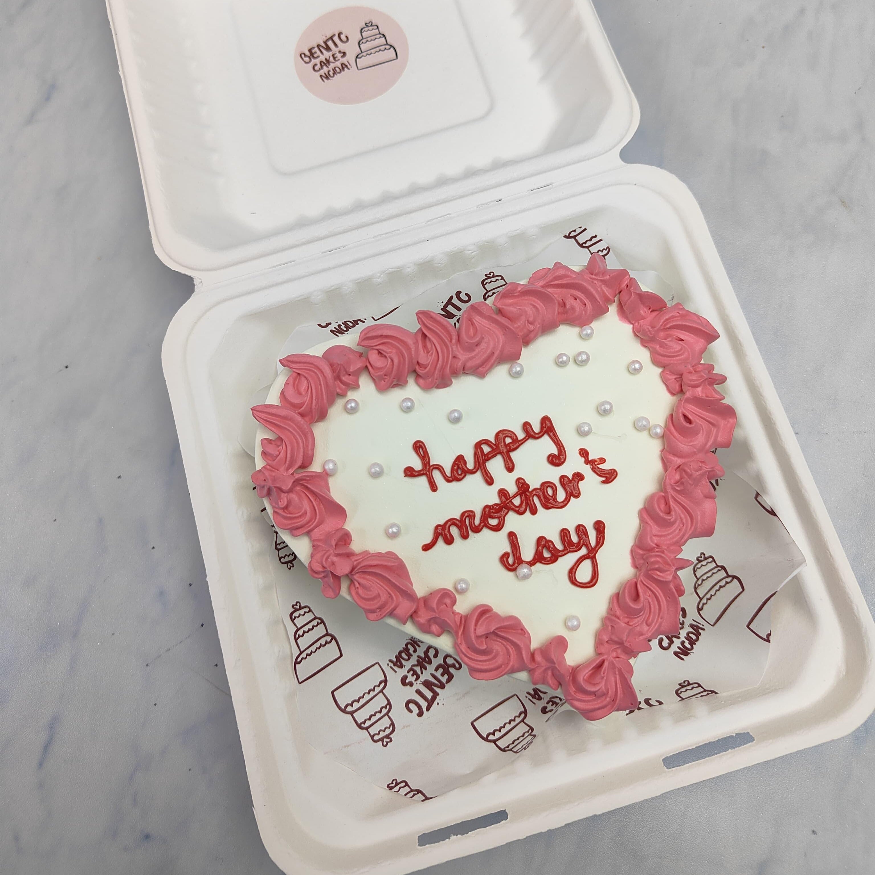 A heart shape white cake with magenta florals design on its border and a "happy mother's day" text written with red icing in its center and decorated with white sprinkles.