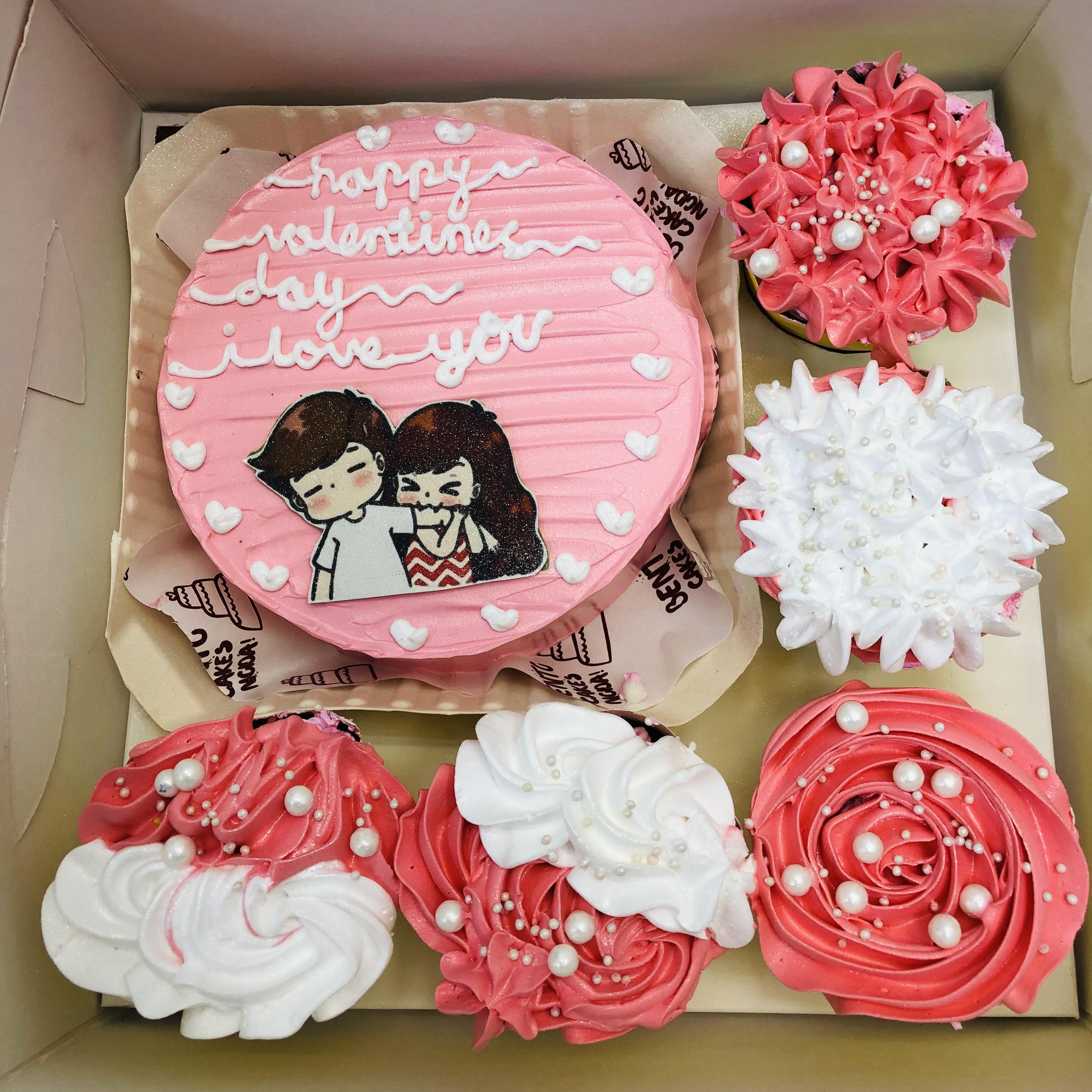 A round white-pink cake and six cupcakes arranged in a bento-style box. The cake has pink frosting with white text that reads "happy valentine's day i love you," along with an illustration of a couple. The cupcakes are decorated with pink and white frosting, some featuring pearl-like decorations.