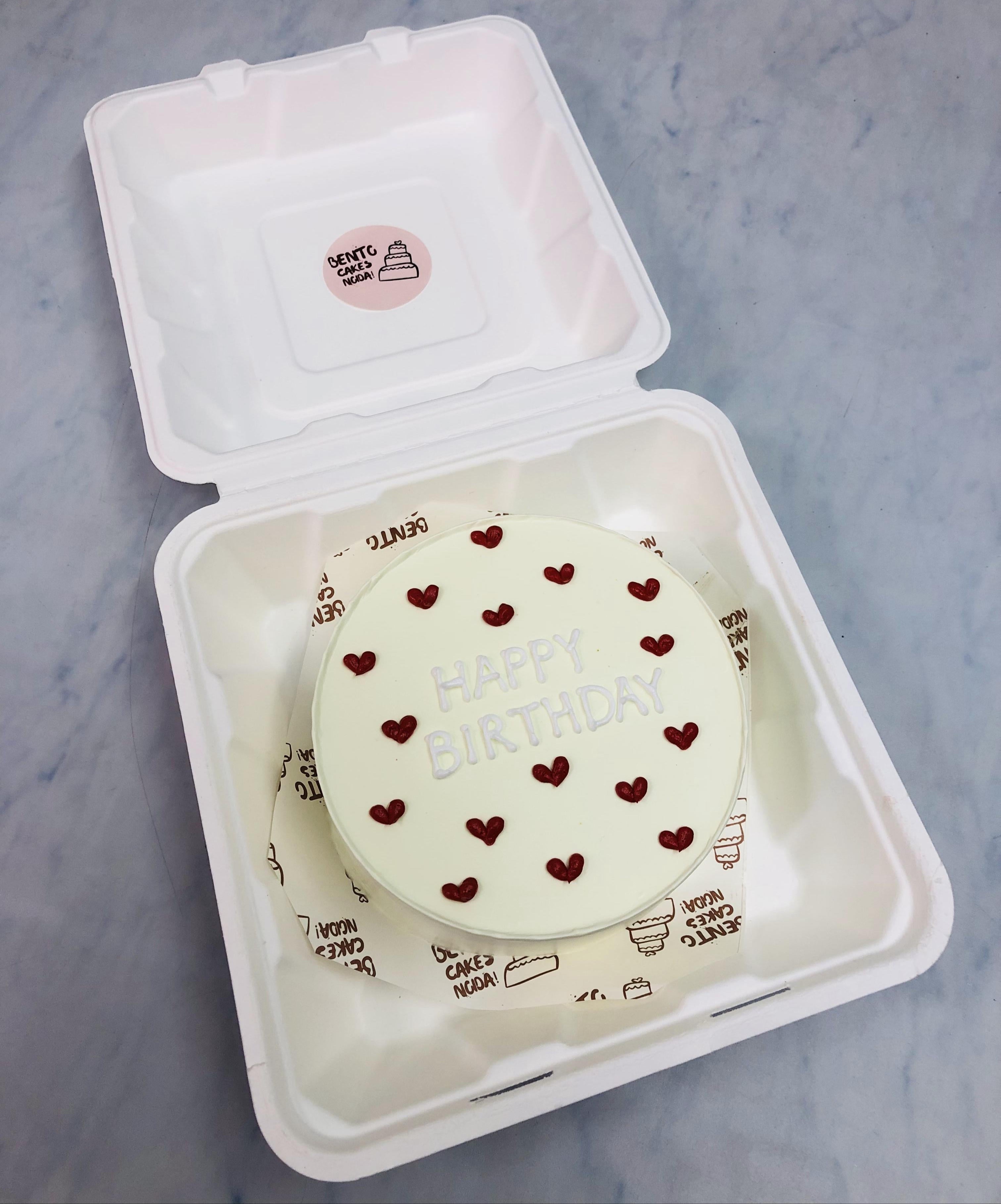 A white bento cake decorated with red hearts and a text "HAPPY BIRTHDAY" written with icing on it.