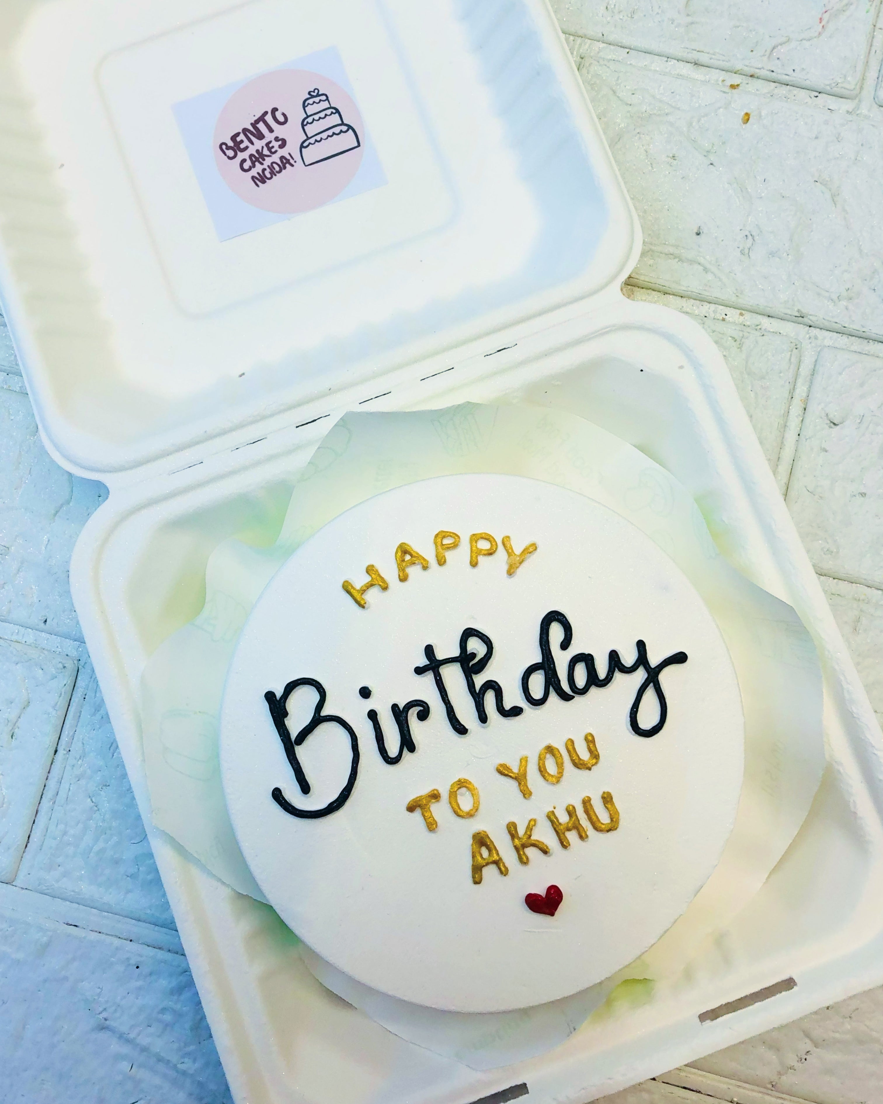 A white bento caqke having golden and black text with a red heart design.