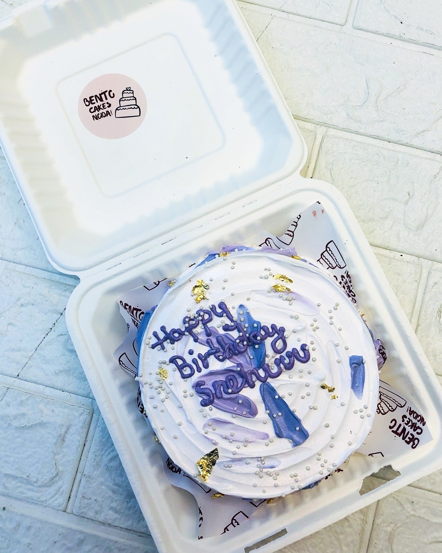 A beautifully decorated bento cake in a white bento box. The cake features rich purple icing, delicate gold leaf accents, white pearls, and a personalized message reading "Happy Birthday Sneha."