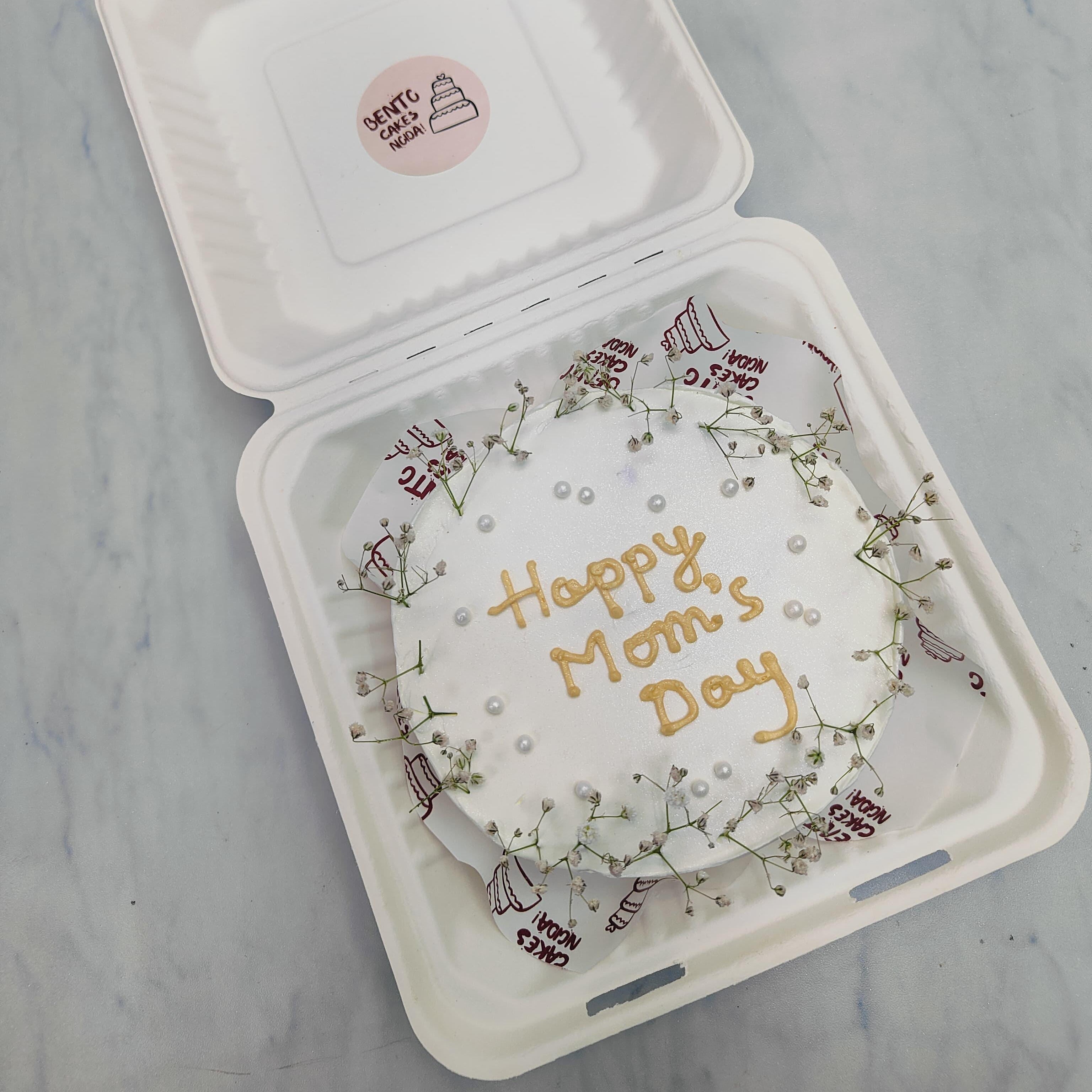 A white cake with aesthetic white florals placed on it's boundry and a text of mother's day is written in it's center.