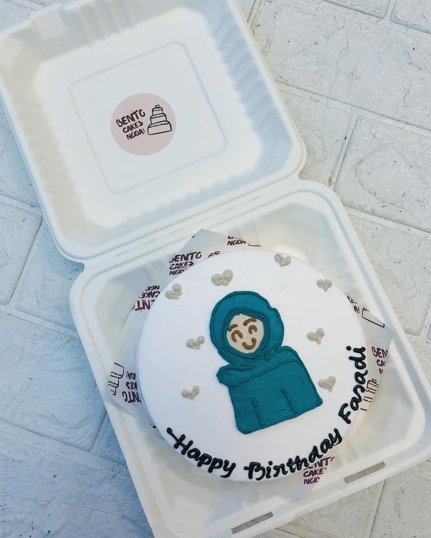 A round birthday cake with white icing, featuring a drawing of a person wearing a teal hijab. The cake is decorated with small heart shapes and the text "Happy Birthday Faradi" written in black icing. 