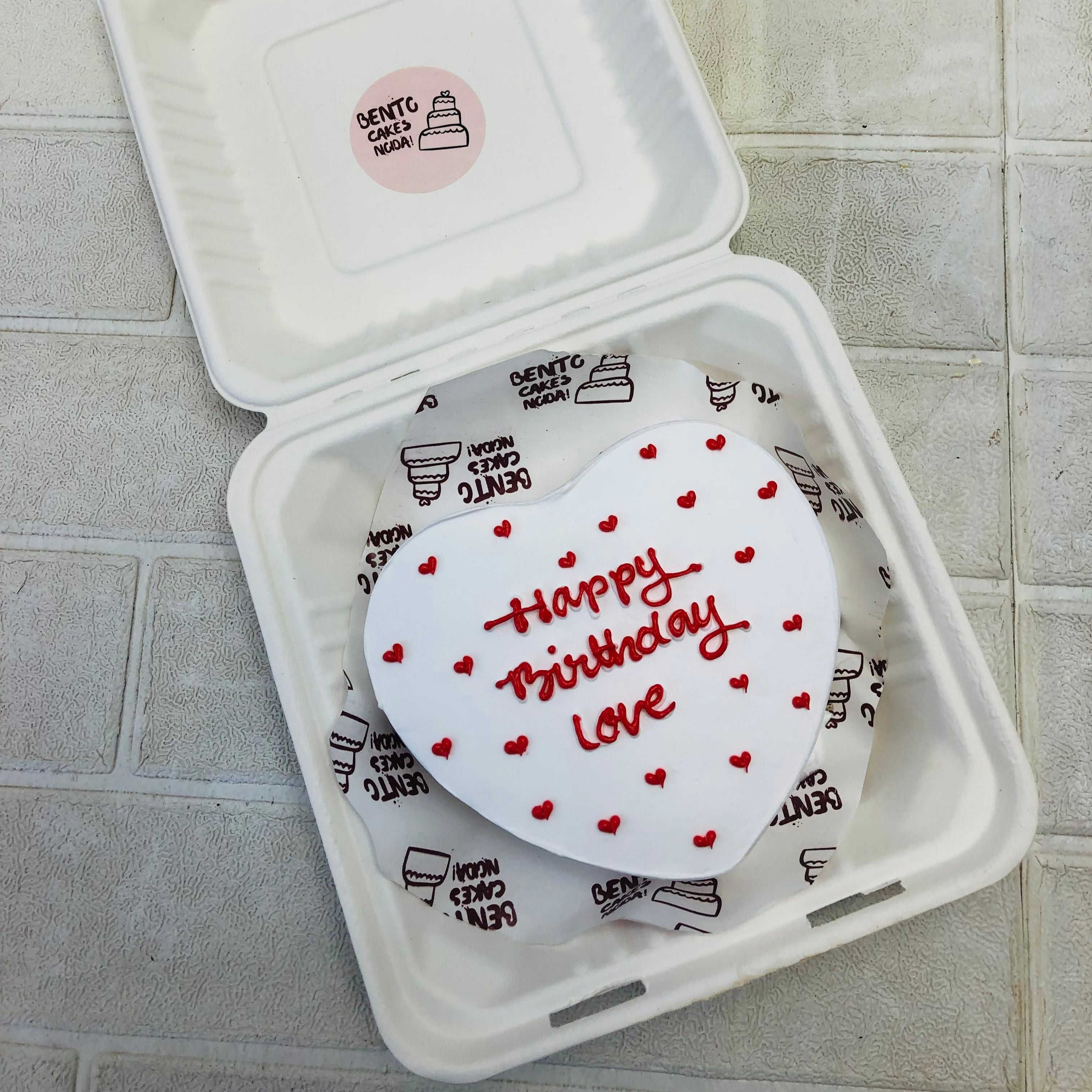 A white icing heart shape cake decorated with little red hearts design and a text "Happy Birthday Love" written with red icing on it.