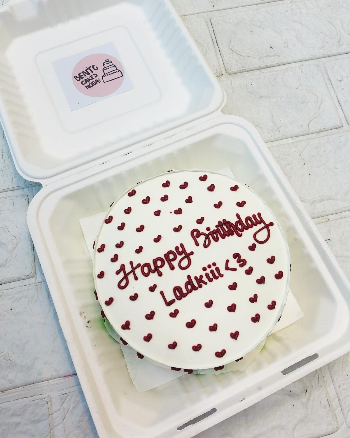 A round birthday cake with smooth white frosting, adorned with delicate red heart decorations. The message "Happy Birthday Ladkiii <3" is written in red icing, adding a personal touch.