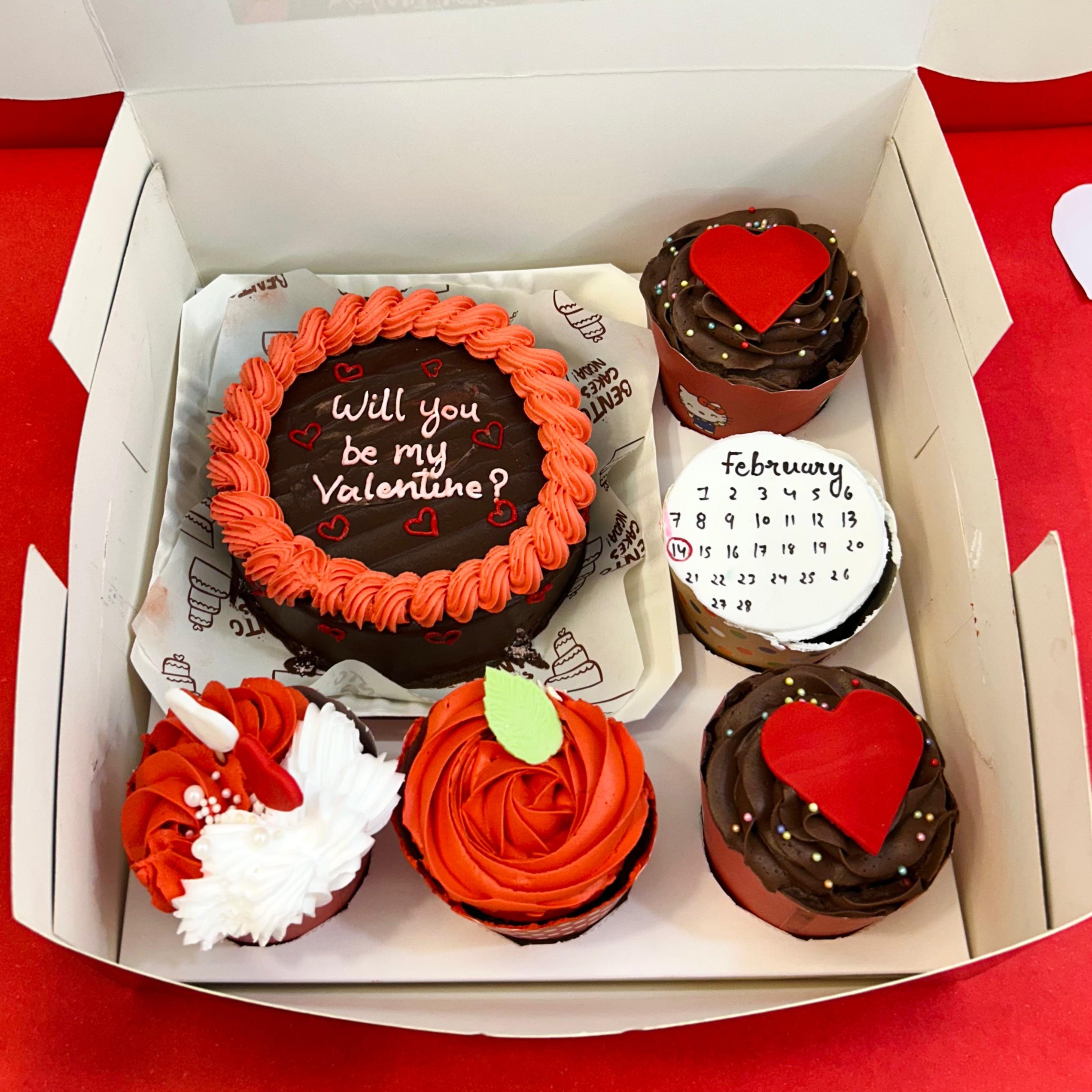 A box of cupcakes featuring a Valentine's Day message: "Will you be my Valentine?" alongside a bento combo cake.