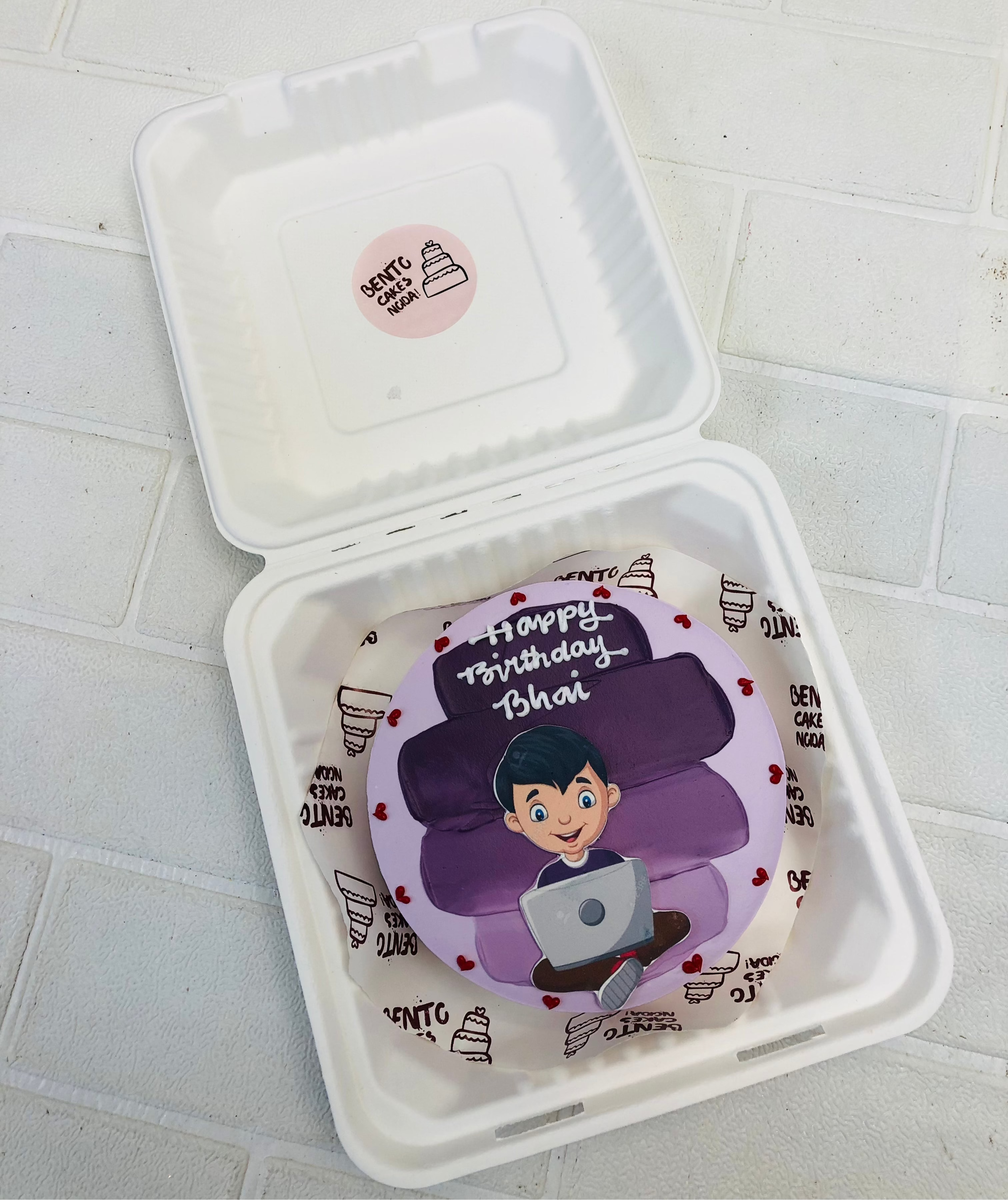 A personalized bento cake in a bento box. The cake is decorated with purple layers and features a figure holding a laptop. The text 'Happy Birthday Bhai' is written on the cake.
