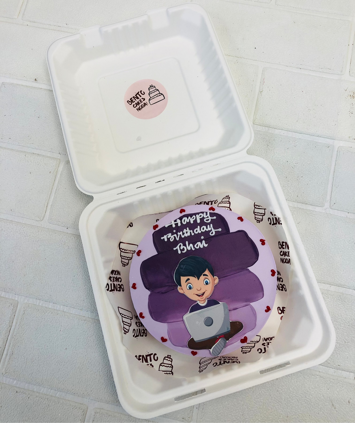 A personalized bento cake in a bento box. The cake is decorated with purple layers and features a figure holding a laptop. The text 'Happy Birthday Bhai' is written on the cake.