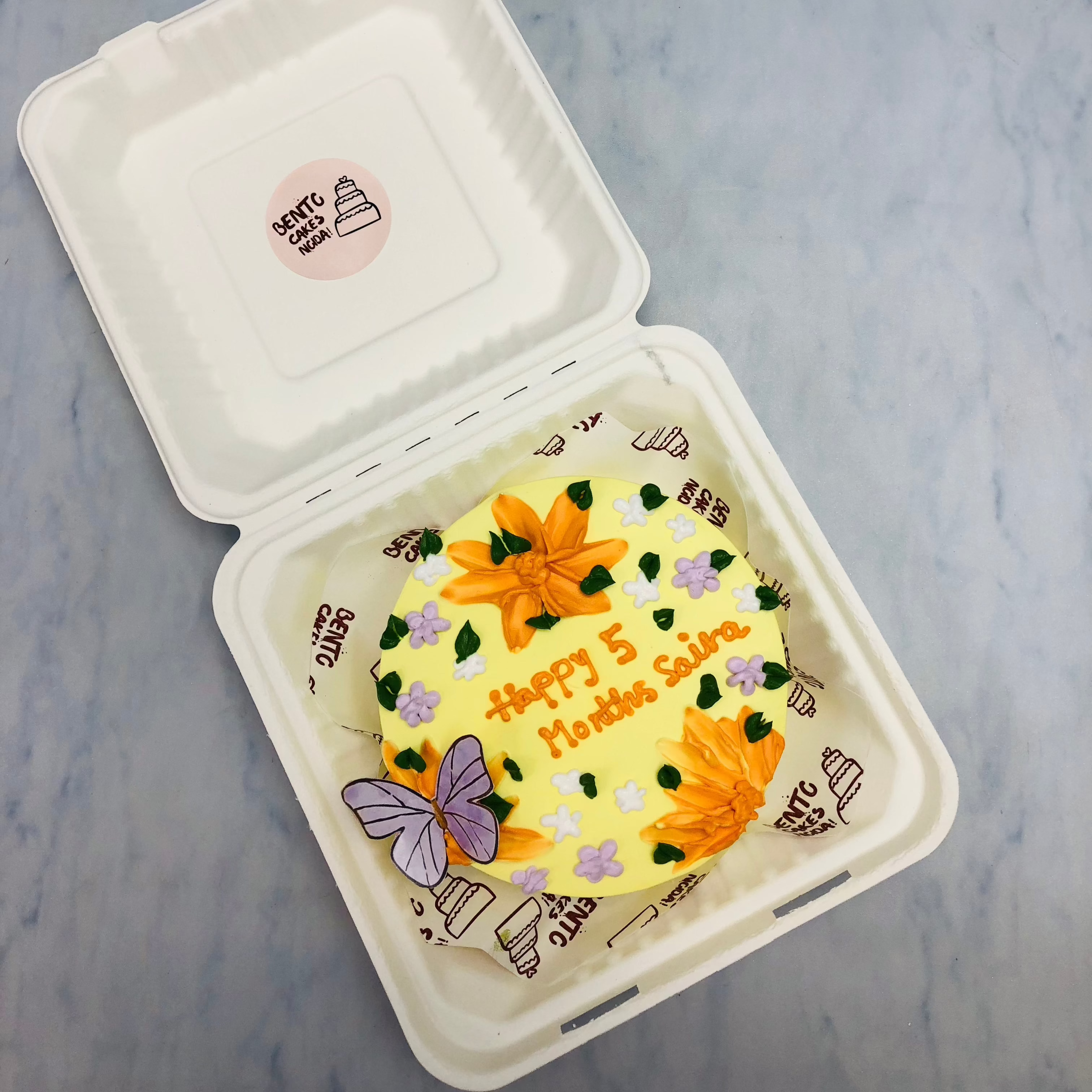A light yellow cake in a bento box for 5 months baby with yellow creamy flower design and a light blue butterfly.