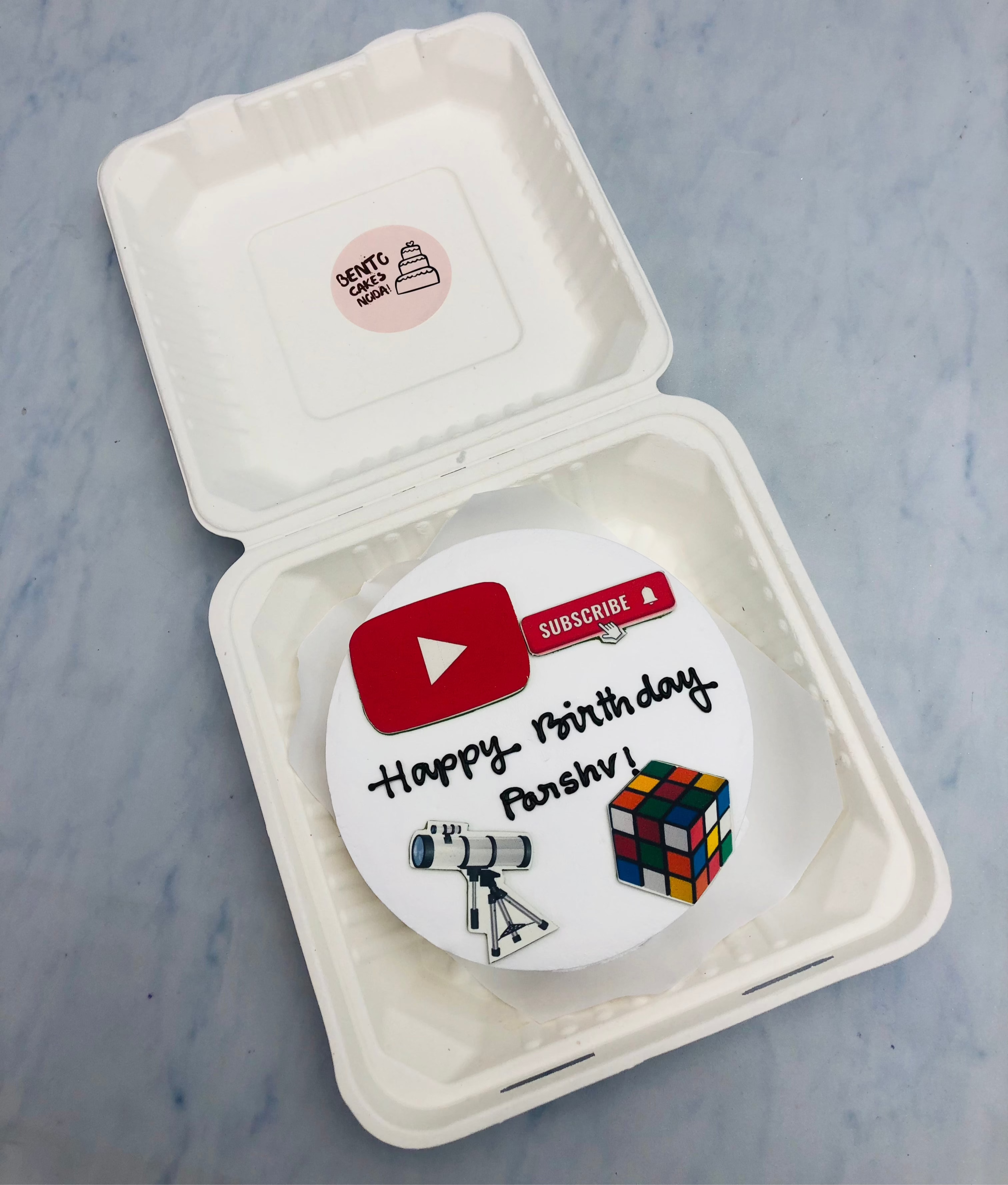 A bento cake with white icing is designed for Youtube subscribers theme with a telescop and puzzle print.