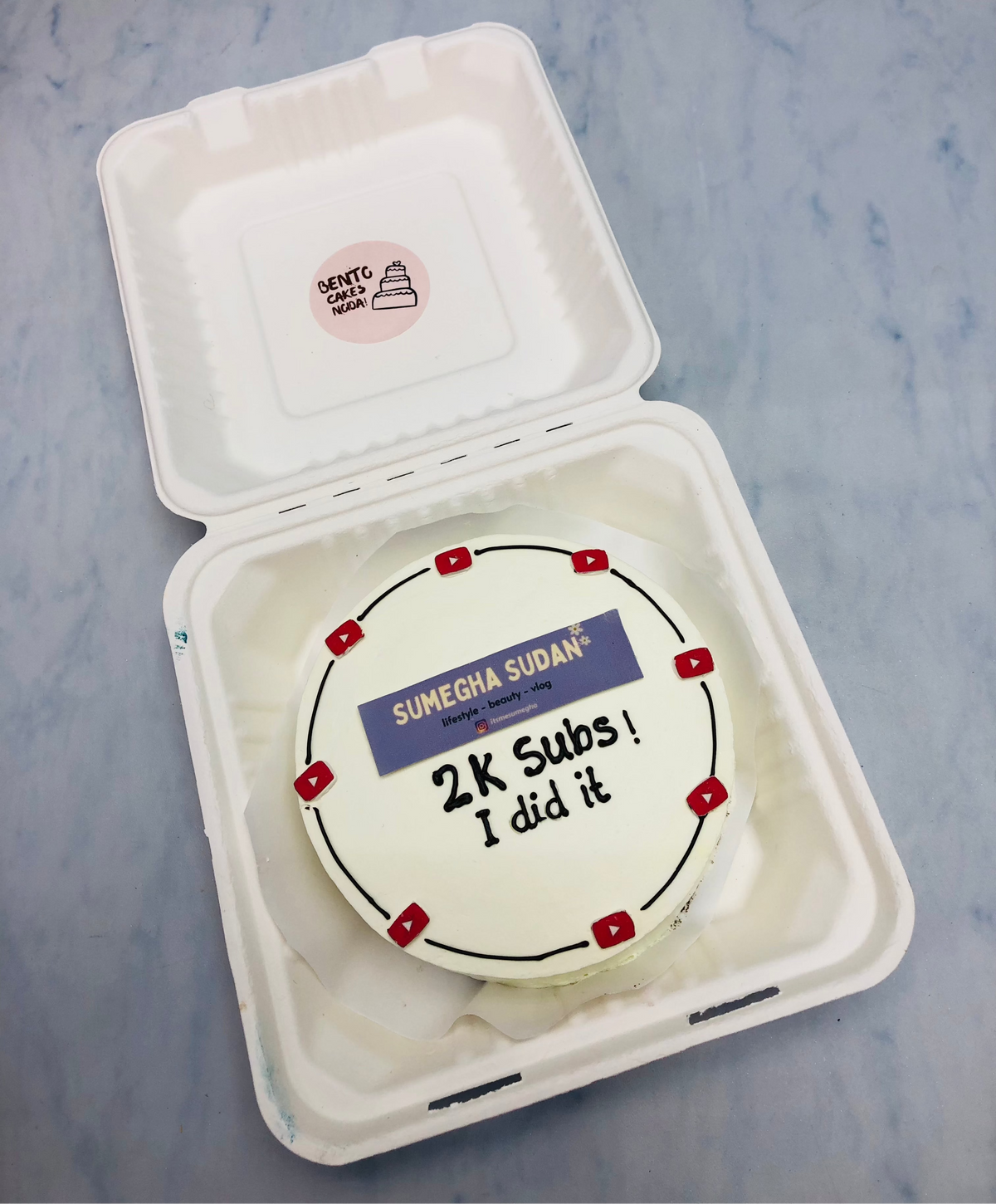 A white icing bento cake designed for 2K subscribers with youtube logo all around the cake.