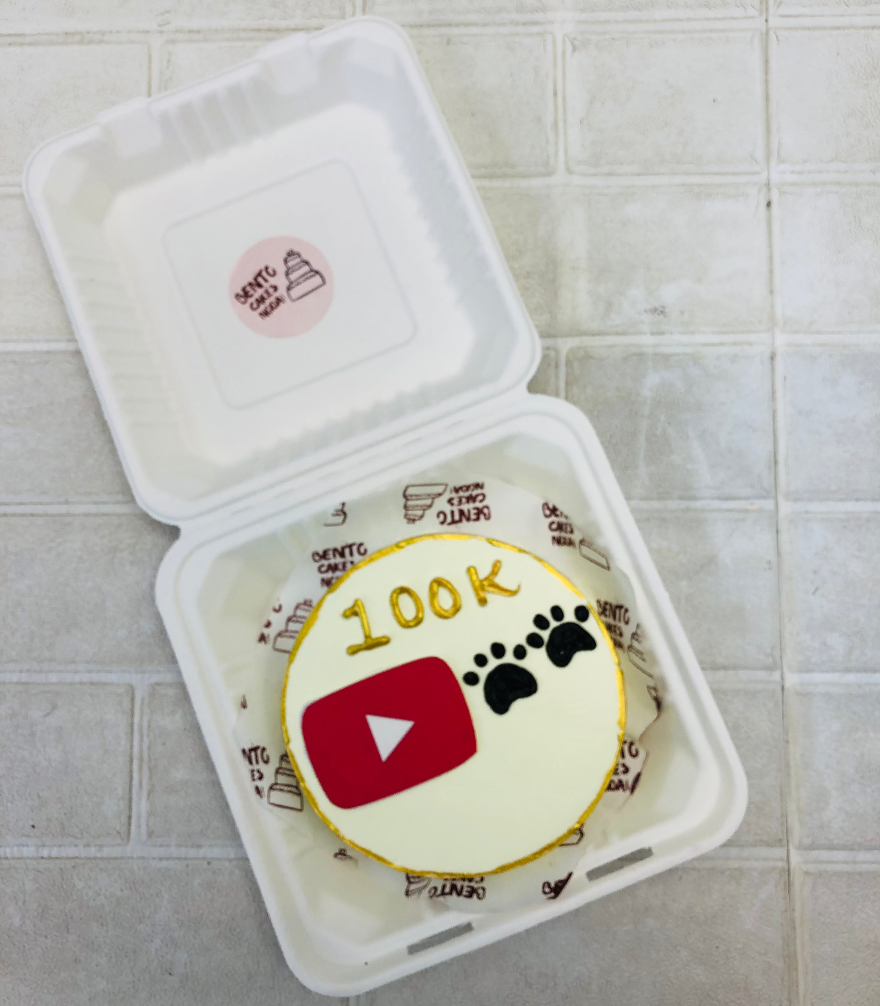 A bento cake is designed with golden bounday and a footprint of puppy with a youtube button logo.