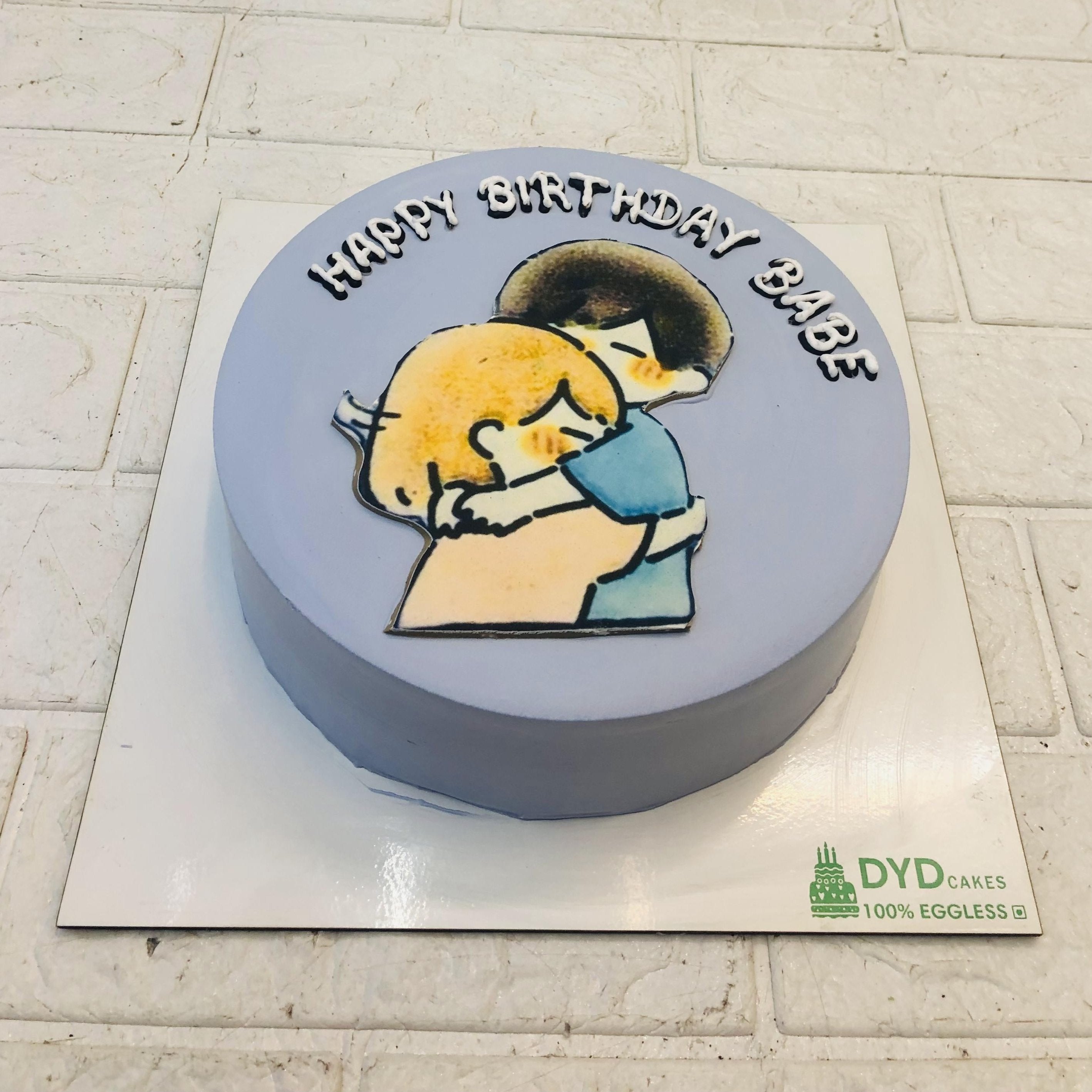 A grey cake having a hugging couple on top with a "HAPPY BIRTHDAY BABE" text written on it.