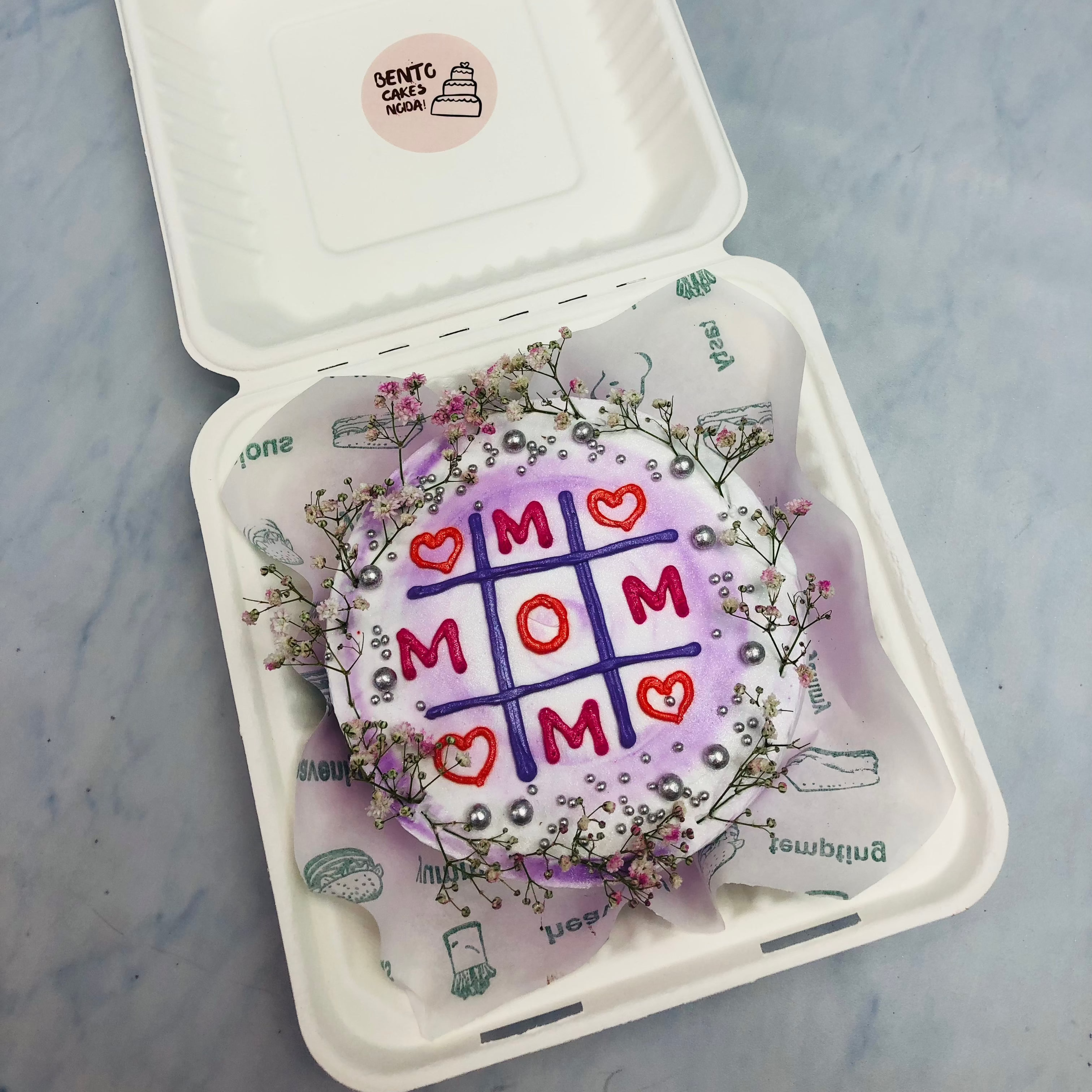An aesthetic floral decorated cake designed with Tik Take Tow game with text "MOM" in it.