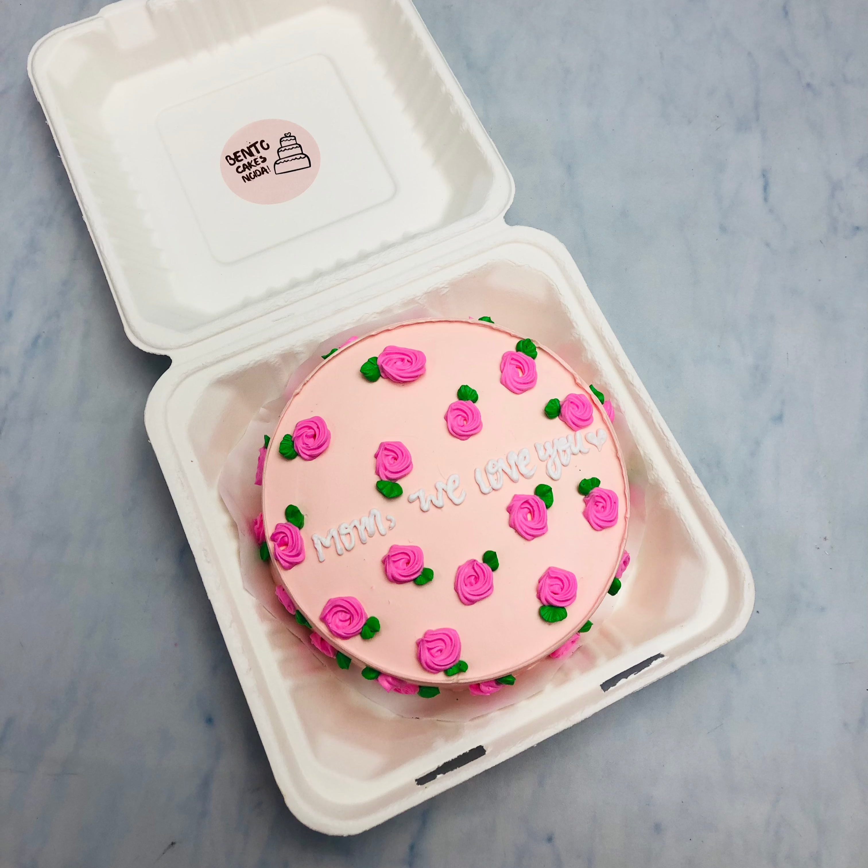 A light pink cake designed with pink floral theme and a text "Mom we love you" beautifully written with white icing.