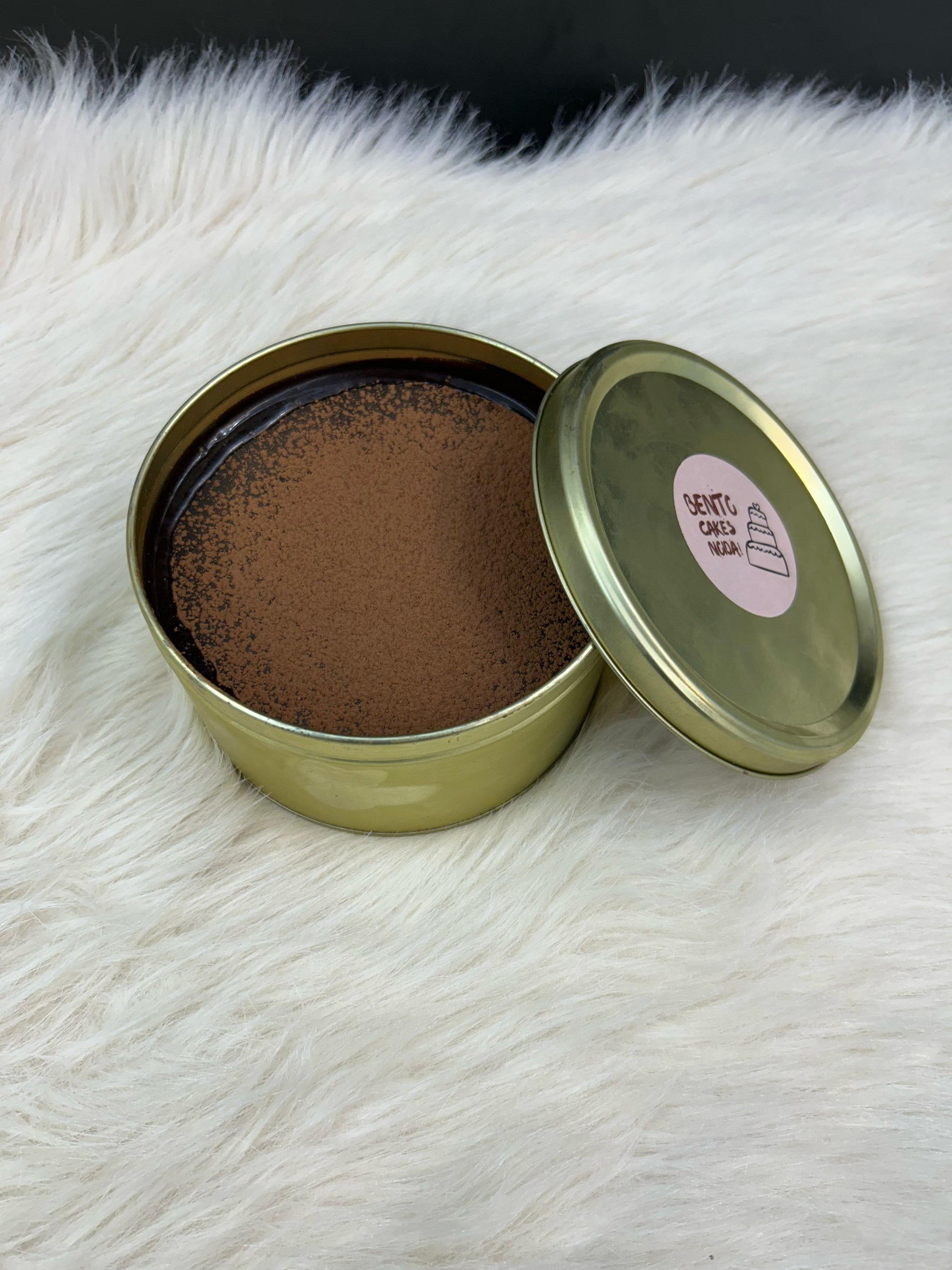 A luxurious Dream Tin Cake enclosed in an elegant golden tin, featuring a cracking chocolate disc, flowing chocolate liquid, luscious cream, and a soft, fluffy cake layer, displayed on a cozy white surface.