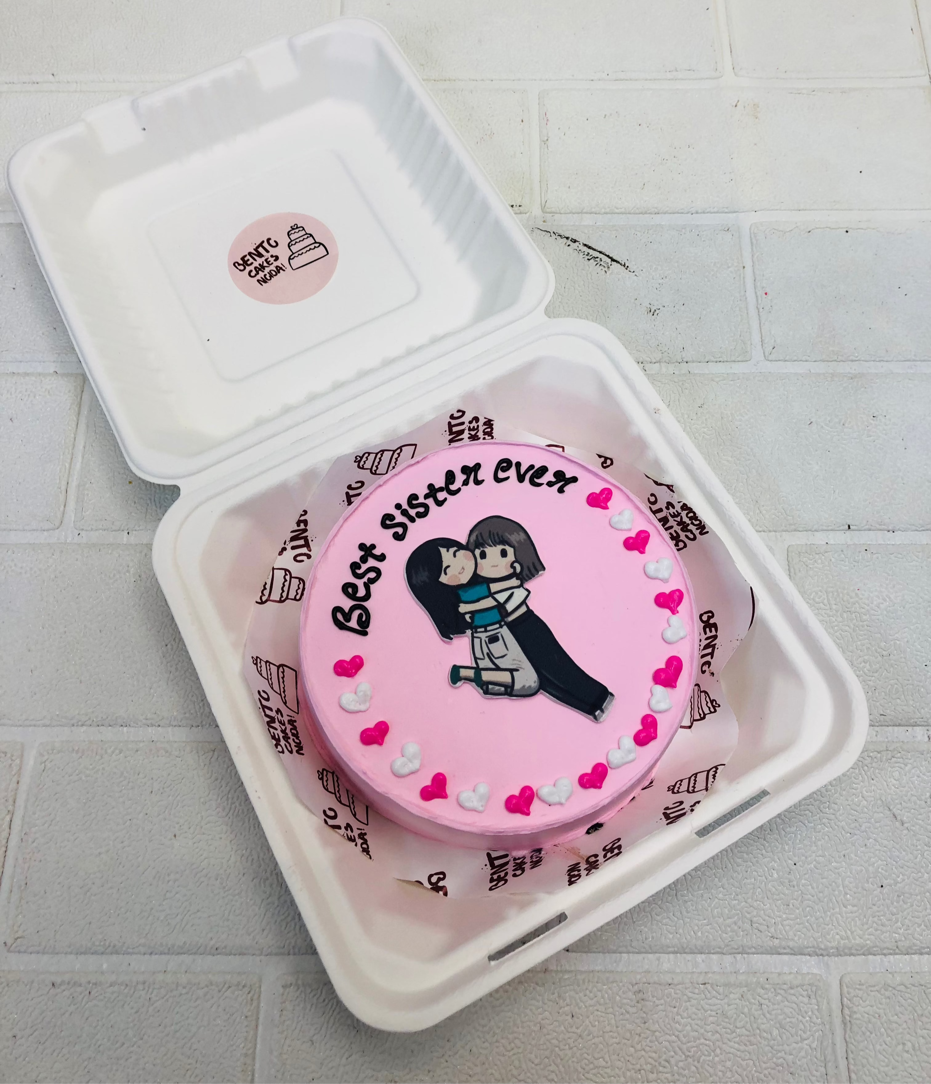 A light pink cake with pink, white hearts design on cake surface and two sisters hugging each ther.